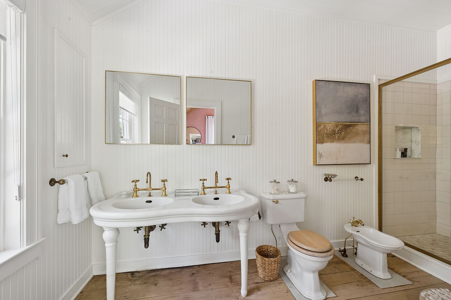 This bright and airy bathroom features a double-sink vanity with brass fixtures and a classic white pedestal base. The space is finished with vertical beadboard wall paneling, light wood flooring, and a walk-in shower enclosed by a brass-framed glass partition. A toilet and bidet are positioned alongside the vanity, while a piece of abstract wall art adds a touch of color to the neutral, elegant aesthetic.