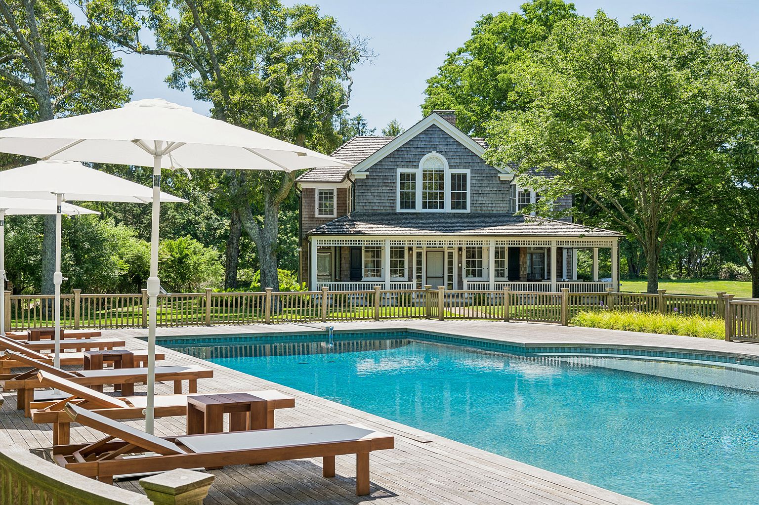 This serene outdoor scene features a large, rectangular swimming pool set within a spacious wooden deck. A row of wooden lounge chairs with white cushions is arranged under white umbrellas, providing a comfortable space for relaxation. In the background, a classic shingle-style house with a covered porch and mature trees creates a picturesque and inviting residential setting.