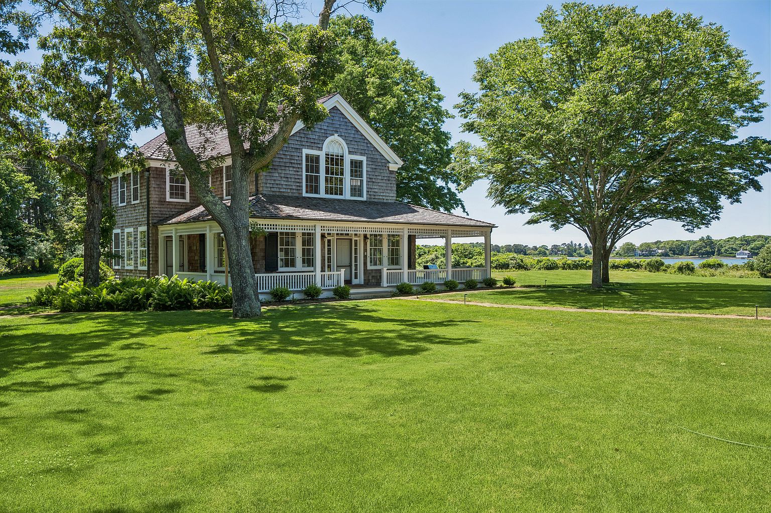 This charming shingle-style home features a wrap-around porch and a classic gabled roofline, set against a backdrop of lush green trees and a serene waterfront. The exterior is clad in weathered-look shingles, complemented by white trim and multi-pane windows that offer a timeless, coastal aesthetic. A sprawling, well-manicured lawn leads up to the residence, emphasizing the property's expansive and peaceful outdoor setting.