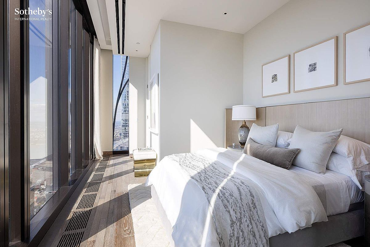 This sophisticated primary bedroom features floor-to-ceiling windows that offer a dramatic, high-rise perspective of the city skyline. The room is elegantly appointed with a plush bed, neutral-toned bedding, and minimalist framed artwork, creating a serene and luxurious atmosphere. The warm sunlight streaming through the glass highlights the rich wood flooring and the clean, modern architectural lines of the space.