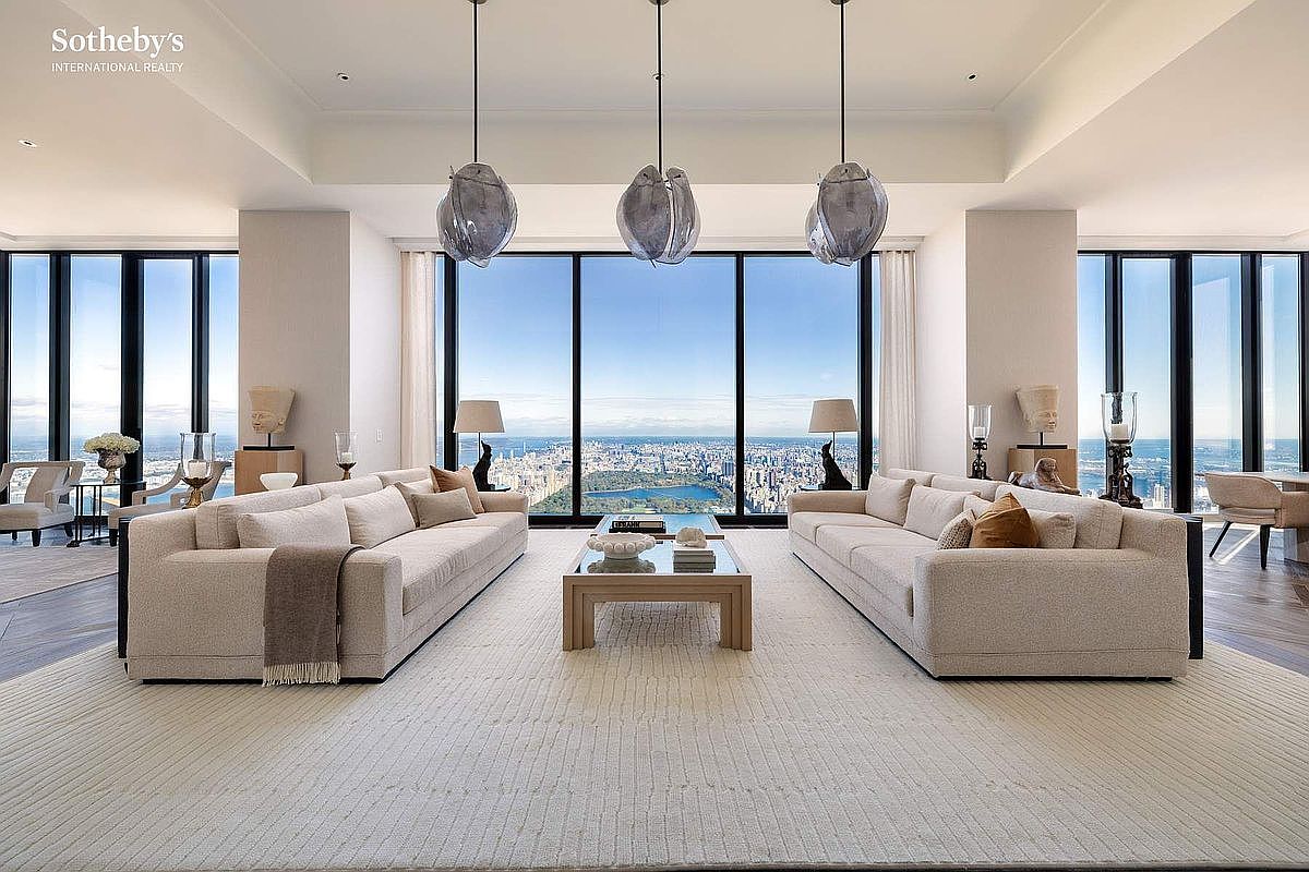 This expansive, high-end living room features a symmetrical layout with two large, plush beige sofas facing each other across a central coffee table. The room is defined by floor-to-ceiling windows that provide a breathtaking, panoramic view of the city skyline and a central park. Three unique, sculptural pendant lights hang from the ceiling, adding a modern artistic touch to the sophisticated and airy space.