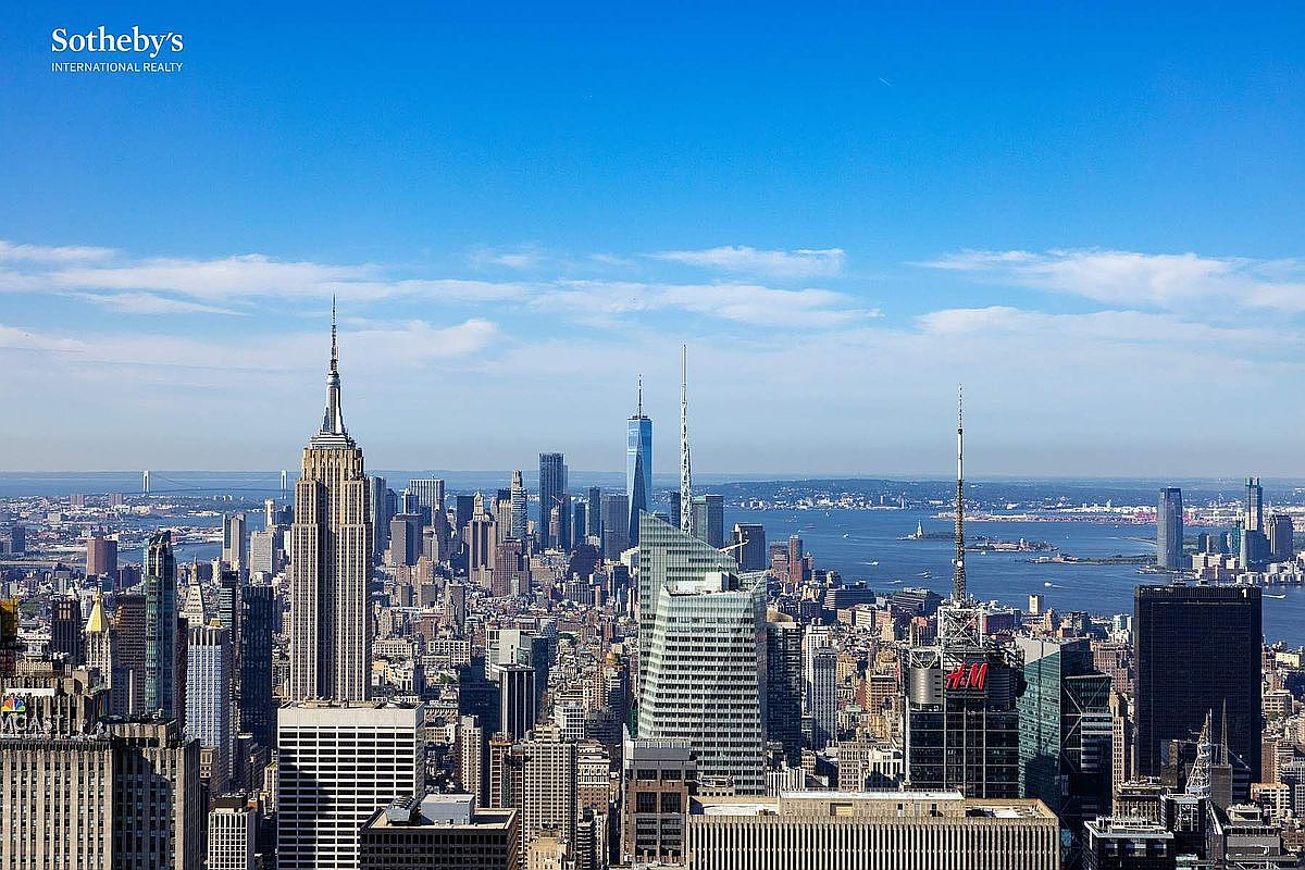 This high-altitude aerial view captures a breathtaking, expansive panorama of the New York City skyline on a clear, sunny day. The iconic Empire State Building stands prominently on the left, while the One World Trade Center is visible in the distance, surrounded by a dense urban landscape of skyscrapers and the shimmering waters of the Hudson River. The perspective offers a cinematic sense of scale and prestige, highlighting the prime location and commanding views that define luxury high-rise living in Manhattan.
