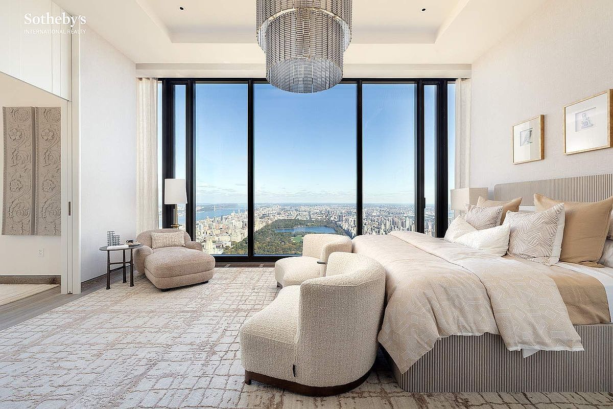 This luxurious primary bedroom features a sophisticated, neutral color palette and floor-to-ceiling windows that offer a breathtaking, panoramic view of Central Park and the New York City skyline. The room is elegantly furnished with a plush, textured bed, curved accent chairs, and a contemporary chandelier, creating an atmosphere of refined urban opulence. The perspective is captured from a slightly elevated eye-level, emphasizing the seamless integration of high-end interior design with the iconic outdoor vista.