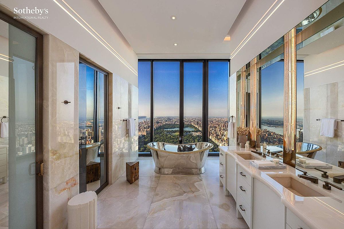 This luxurious primary bathroom features a stunning metallic soaking tub positioned perfectly in front of floor-to-ceiling windows that offer a breathtaking panoramic view of Central Park and the New York City skyline. The space is finished with elegant marble surfaces, dual vanity sinks, and sophisticated lighting, creating an atmosphere of opulence and high-end urban living. The perspective is centered, drawing the eye directly toward the dramatic cityscape beyond the glass.