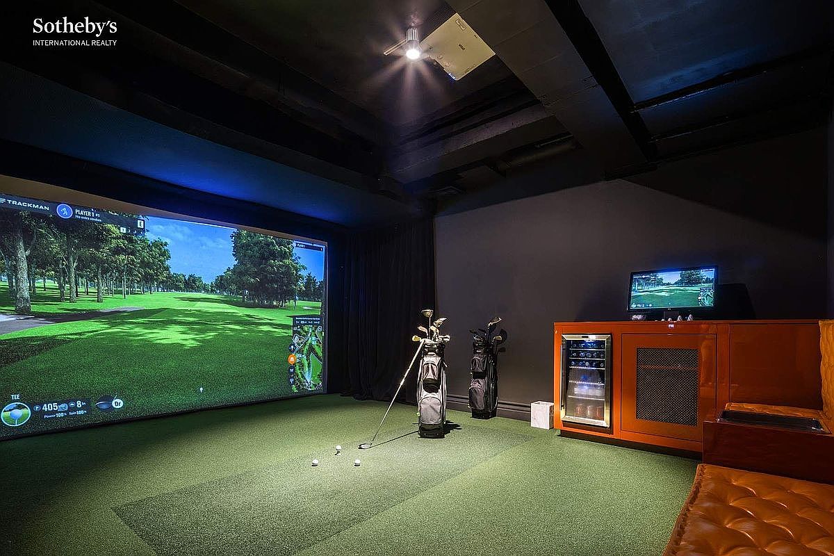 This image showcases a high-end home golf simulator room, featuring a large projection screen displaying a virtual golf course and a synthetic turf floor. The space is equipped with golf bags, a club, and a sleek orange cabinet unit that houses a beverage cooler and a computer monitor for tracking performance. The room is designed with a dark, moody aesthetic, creating an immersive and professional environment for indoor sports.