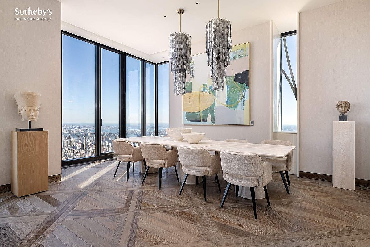 This sophisticated dining room features a long, light-toned stone table surrounded by six upholstered chairs, set against a backdrop of floor-to-ceiling windows offering a dramatic city skyline view. Two ornate, tiered crystal chandeliers hang above the table, complementing the abstract artwork on the wall and the intricate herringbone wood flooring. The space exudes a luxurious, modern aesthetic with a neutral color palette and curated sculptural accents.