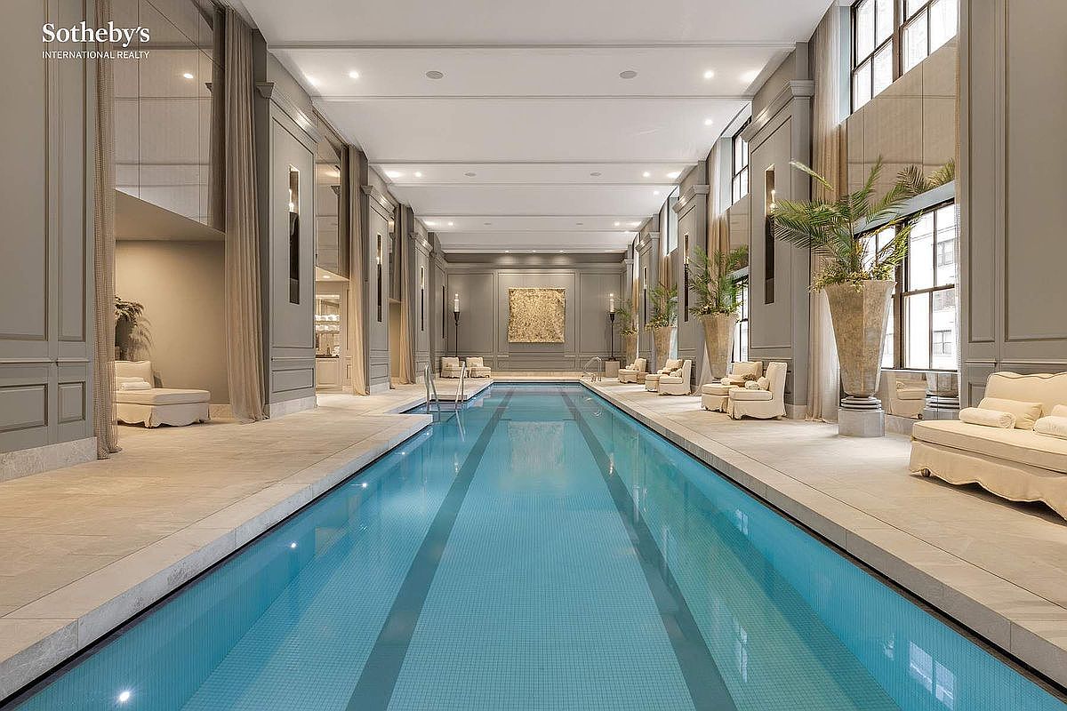 This luxurious indoor lap pool area exudes elegance with its symmetrical design, high ceilings, and sophisticated gray paneling. The space is lined with plush lounge seating, large potted palms, and a central piece of textured wall art, creating a serene, spa-like atmosphere. The perspective is a centered, eye-level shot that emphasizes the length and grandeur of the pool, highlighting its high-end, resort-style aesthetic.