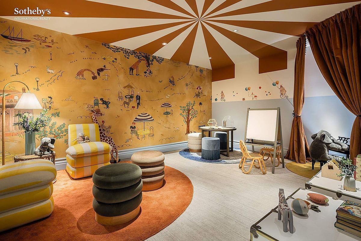 This whimsical children's playroom features a vibrant circus-tent-inspired ceiling and a charming illustrated wallpaper depicting a playful outdoor scene. The room is furnished with colorful, plush seating, a small activity table, and a child-sized easel, creating an inviting and imaginative space. The perspective is a wide-angle shot that captures the entire room's playful decor and cozy, well-appointed layout.