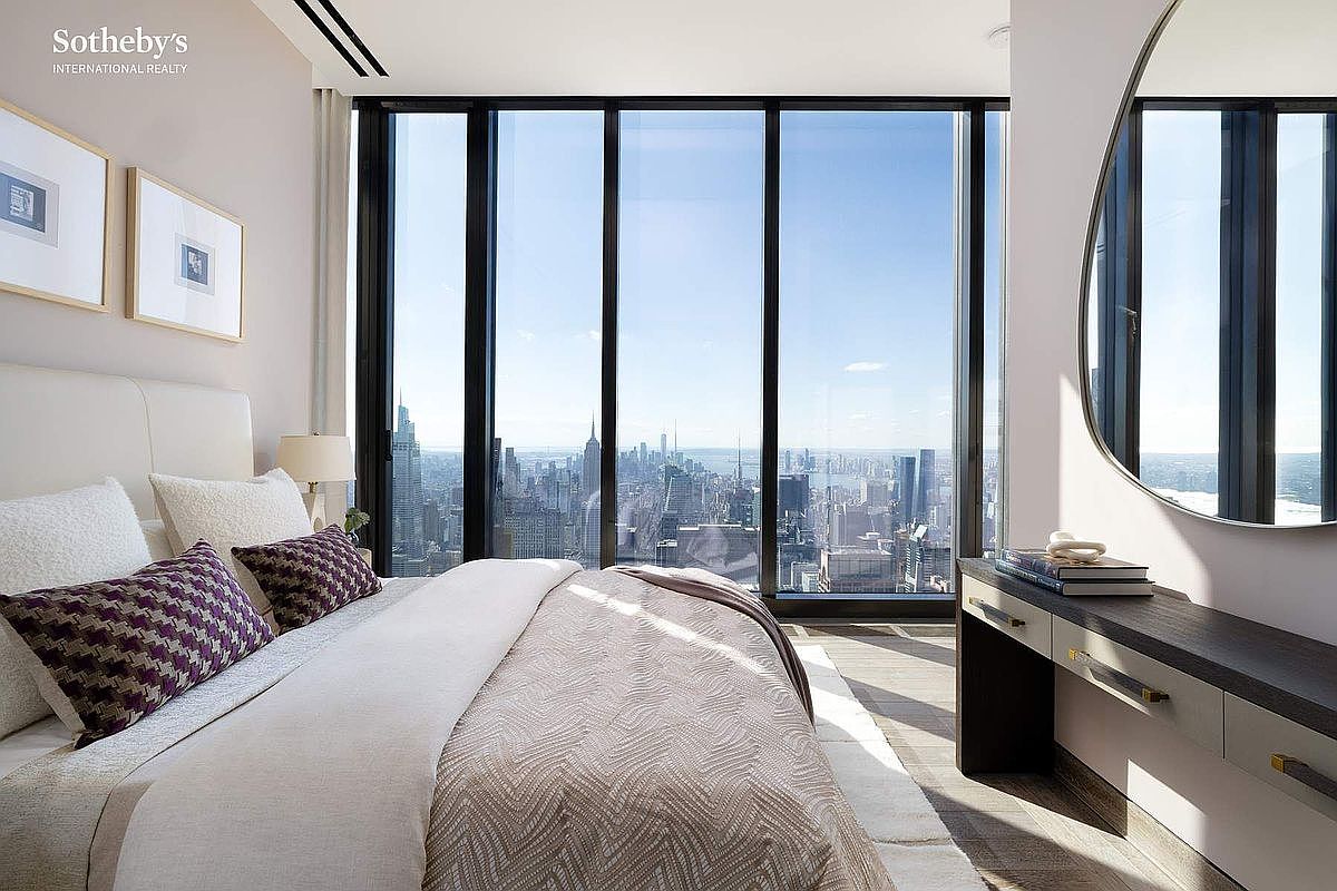 This sophisticated primary bedroom features a plush bed adorned with patterned purple throw pillows and a neutral-toned duvet, set against a backdrop of floor-to-ceiling windows that offer a breathtaking, panoramic view of the New York City skyline. To the right, a sleek, modern vanity with a large circular mirror provides a functional yet elegant touch, while framed artwork adds a refined aesthetic to the light-colored walls. The room exudes a sense of high-end luxury and tranquility, perfectly framing the urban landscape as a central design element.