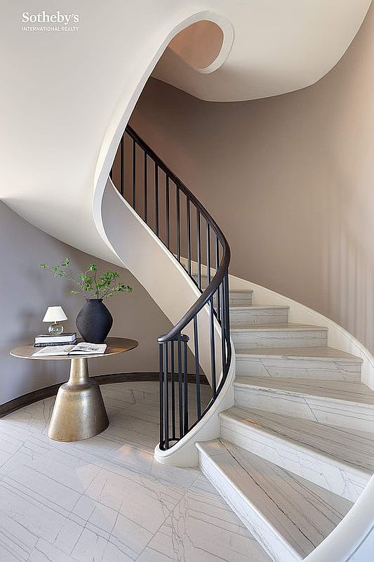 This elegant entryway features a dramatic, sweeping curved staircase with white marble steps and a sleek black metal railing. To the side, a sophisticated gold-toned pedestal table holds a minimalist vase with greenery and a small lamp, creating a welcoming focal point. The space is defined by its clean, modern aesthetic, neutral color palette, and high-end architectural curves.