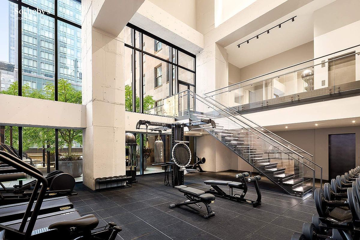 This high-end, multi-level home gym features floor-to-ceiling windows that flood the space with natural light and offer an expansive view of the urban surroundings. The room is equipped with professional-grade fitness equipment, including a central functional training rig, weight benches, and a rack of dumbbells, all set against a sleek, modern architectural backdrop with a glass-railed staircase. The industrial-chic aesthetic, characterized by exposed concrete columns and dark flooring, creates a sophisticated and motivating environment for exercise.