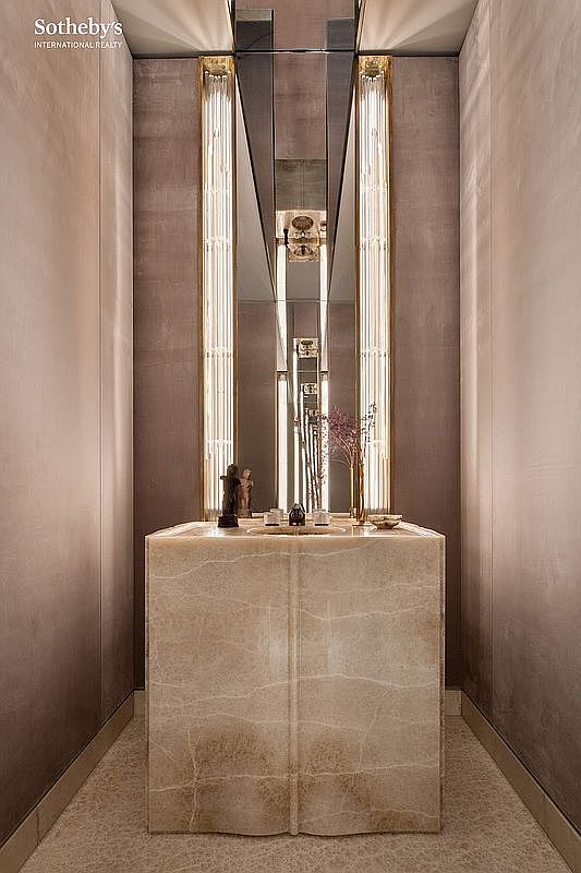This sophisticated powder room features a striking, monolithic stone vanity with a textured, marble-like finish that serves as the room's centerpiece. Flanking the central mirror are two vertical, illuminated light fixtures that cast a warm, elegant glow, enhancing the luxurious atmosphere. The space is framed by smooth, neutral-toned walls and a subtle, patterned floor, creating a refined and intimate aesthetic.