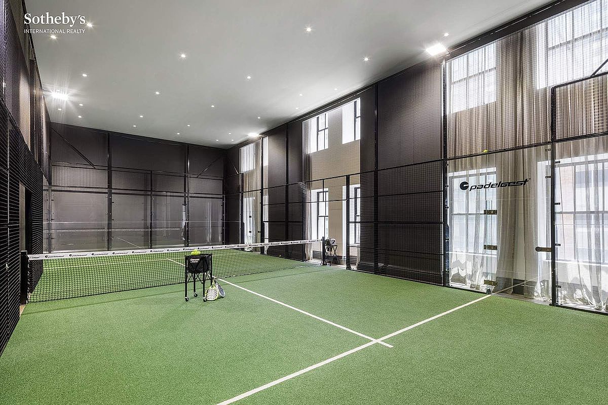 This image showcases a high-end, indoor padel court featuring professional-grade green turf, a central net, and a ball hopper with rackets. The space is enclosed by black metal fencing and glass panels, set within a modern, high-ceilinged room with recessed lighting and large windows draped in sheer curtains. The overall aesthetic is sleek, athletic, and luxurious, highlighting a unique recreational amenity within a residential property.