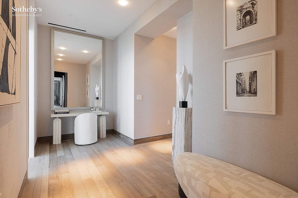 This elegant entryway or hallway features light-toned hardwood flooring and textured neutral wallpaper, creating a sophisticated and airy atmosphere. A sleek console table with a large mirror serves as a focal point, complemented by a sculptural art piece on a stone pedestal and framed black-and-white photography on the wall. The perspective is a wide-angle view looking down the corridor, emphasizing the clean lines and high-end, minimalist design aesthetic.