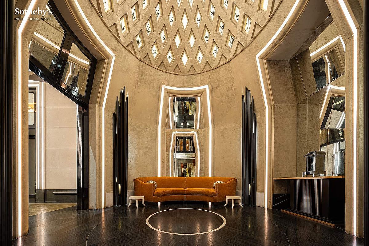 This grand, Art Deco-inspired foyer features a dramatic vaulted ceiling with a geometric lattice skylight and striking vertical LED accent lighting. A plush, burnt-orange sofa serves as a focal point against the textured, warm-toned walls, while a polished dark wood floor with a circular inlay anchors the space. The symmetrical design and sophisticated lighting create an atmosphere of opulent, cinematic luxury.