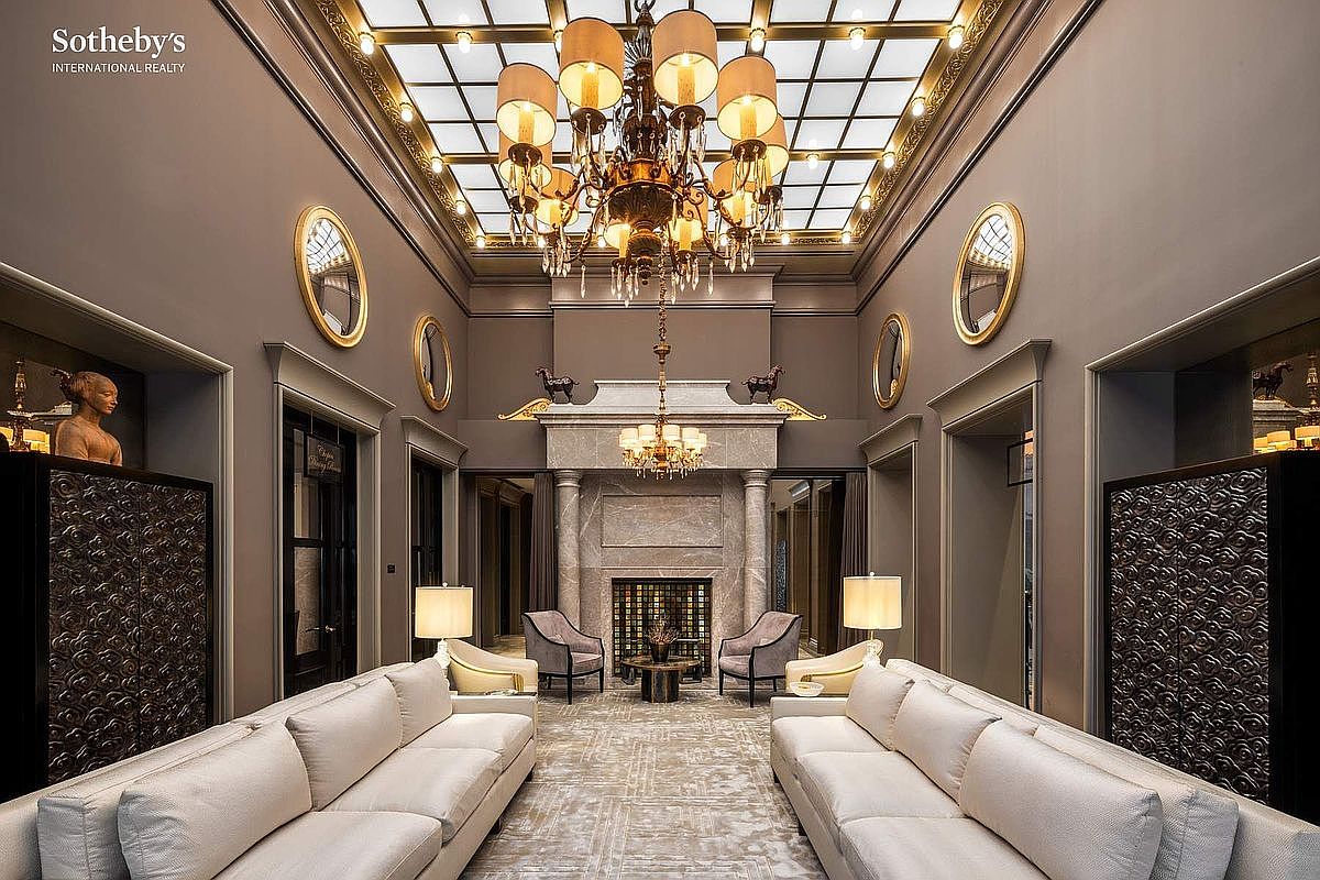 This grand, symmetrical living room exudes opulence with its soaring ceiling featuring a large glass skylight and an ornate, multi-tiered chandelier. The space is anchored by a massive stone fireplace flanked by two armchairs, while two long, cream-colored sofas face each other to create a formal seating arrangement. Elegant wall sconces, decorative mirrors, and intricate metal screens add to the sophisticated, high-end aesthetic of this luxurious interior.
