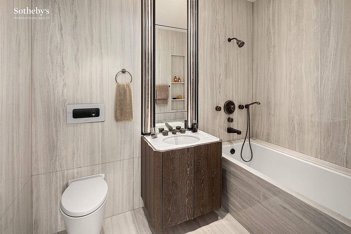 This modern, compact bathroom features elegant, light-toned stone wall tiling and a sleek, dark wood vanity with a white marble countertop. The space is anchored by a wall-mounted toilet and a deep soaking tub, complemented by a tall, framed mirror that enhances the room's verticality. The overall aesthetic is sophisticated and minimalist, creating a luxurious feel within a small footprint.