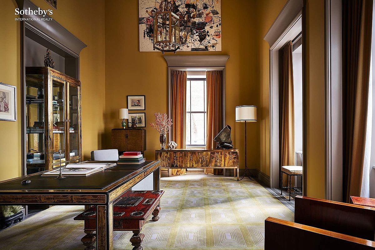 This sophisticated home office exudes a refined, classic aesthetic with its rich mustard-toned walls and elegant, dark wood furnishings. The room features a prominent executive desk, a decorative display cabinet, and a geometric-patterned area rug, all illuminated by a grand chandelier and natural light from a tall window. The perspective is a wide, eye-level shot that captures the balanced, luxurious atmosphere of a high-end study.
