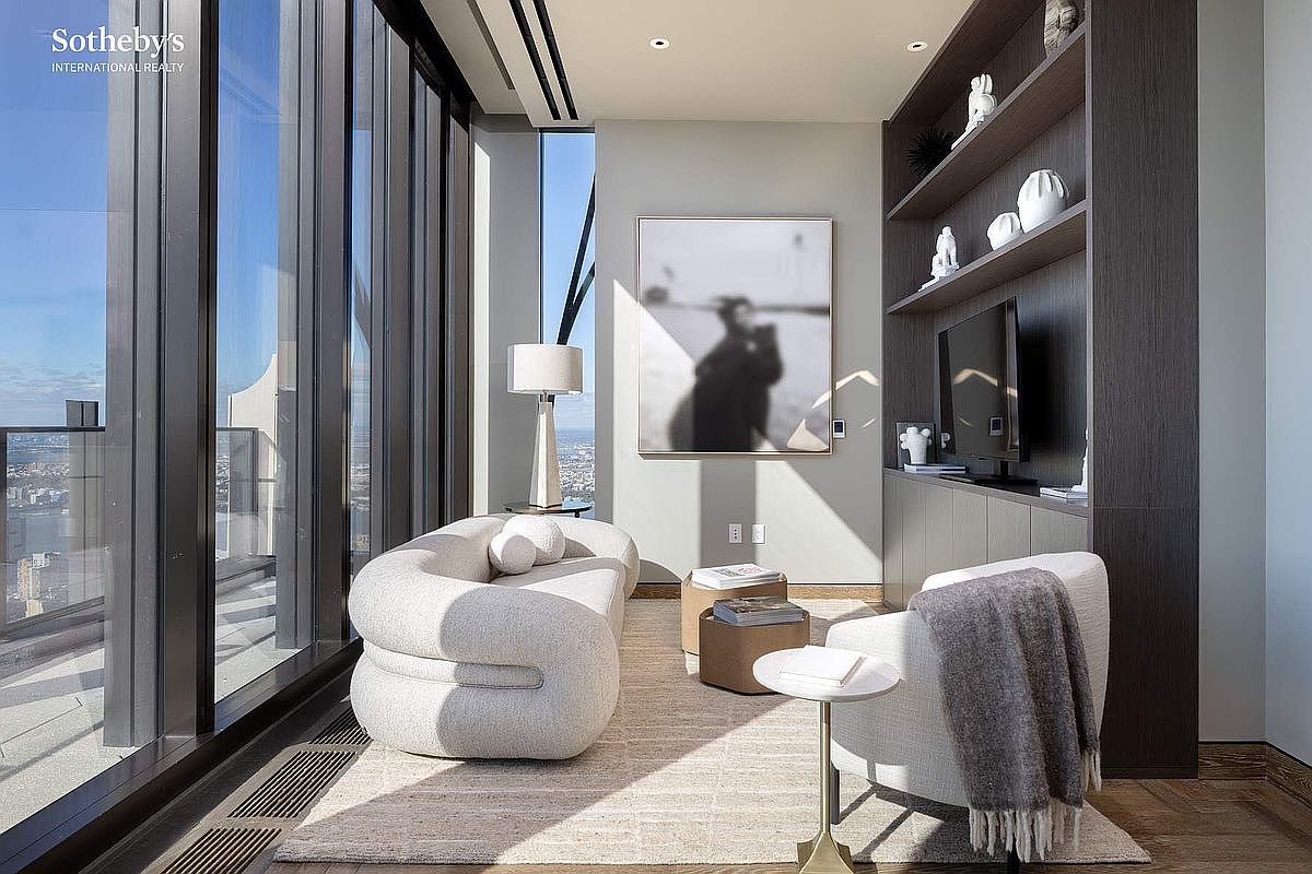 This sophisticated, modern living space features a curved, textured sofa and a matching armchair arranged on a neutral area rug, creating an inviting conversation area. A large, abstract black-and-white artwork serves as a focal point on the wall, complemented by custom dark wood shelving and a sleek television. Floor-to-ceiling windows provide an expansive, cinematic view of the city skyline, bathing the room in natural light and emphasizing the high-rise luxury aesthetic.