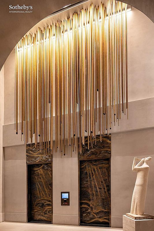 This sophisticated elevator lobby features a dramatic, floor-to-ceiling installation of vertical gold-toned metallic rods that create a cascading, light-catching effect. The two elevator doors are finished in a rich, swirling dark marble or stone, providing a striking contrast to the minimalist, light-colored walls. A sculptural figure stands on a pedestal to the right, adding an artistic and refined touch to this high-end residential or commercial entryway.