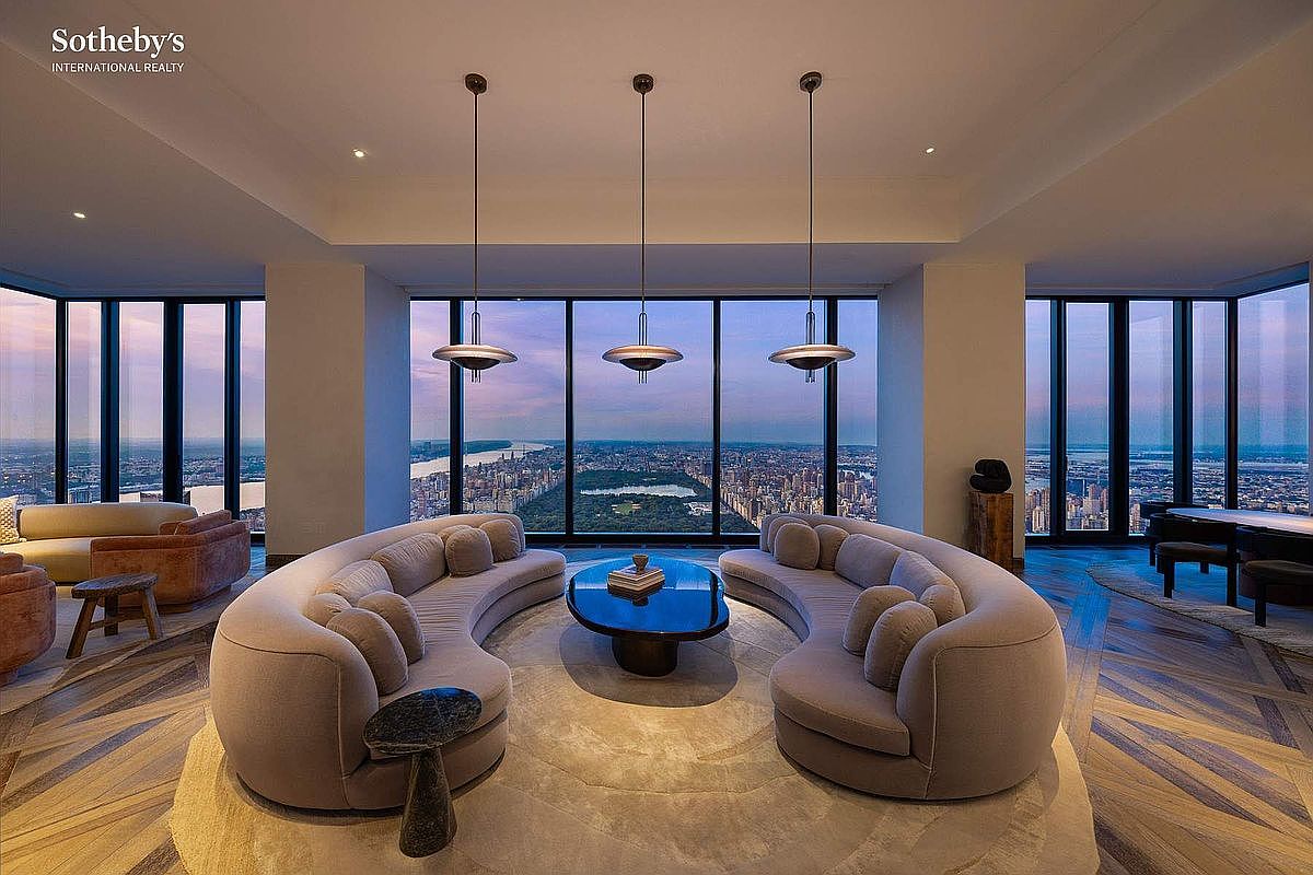 This luxurious, high-rise living room features a sophisticated, symmetrical layout centered around two curved, plush sofas facing a dark, oval coffee table. Floor-to-ceiling windows provide a breathtaking, panoramic view of the city skyline and Central Park at dusk, creating a dramatic and opulent atmosphere. The room is accented by three sleek, modern pendant lights and rich, patterned hardwood flooring, emphasizing a refined and contemporary aesthetic.