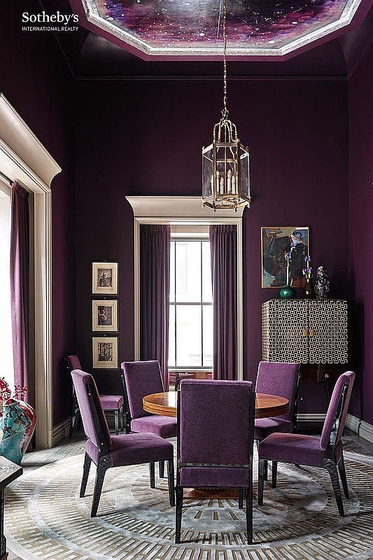 This sophisticated dining room features deep purple walls and matching upholstered chairs arranged around a circular wooden table. A striking, ornate lantern-style chandelier hangs from a ceiling painted with a celestial, nebula-inspired mural, while a patterned cabinet and framed artwork add elegant detail. The room exudes a luxurious and dramatic atmosphere, perfectly framed by tall, dark curtains and architectural molding.