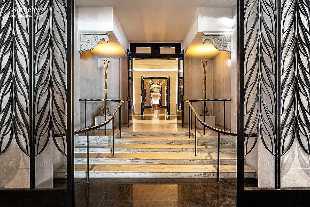 This grand, symmetrical entryway features a set of marble stairs leading up to a secondary doorway, framed by ornate, leaf-patterned metal screens. The space is accented by decorative stone corbels, warm recessed lighting, and tall, slender pedestals topped with sculptural elements. The perspective is centered and cinematic, emphasizing a sense of luxury, formal elegance, and architectural depth.
