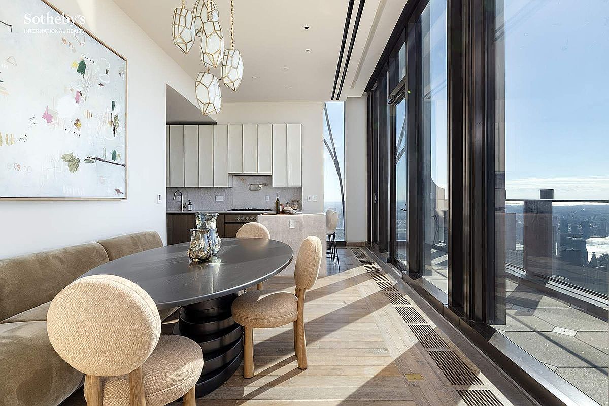 This sophisticated dining area features a sleek, dark oval table paired with modern, light-toned upholstered chairs, set against a backdrop of floor-to-ceiling windows that offer expansive city views. A large, abstract piece of art hangs on the wall, while unique geometric pendant lights provide a focal point above the table. The open-concept space seamlessly transitions into a minimalist kitchen, creating an airy and luxurious atmosphere perfect for high-end urban living.