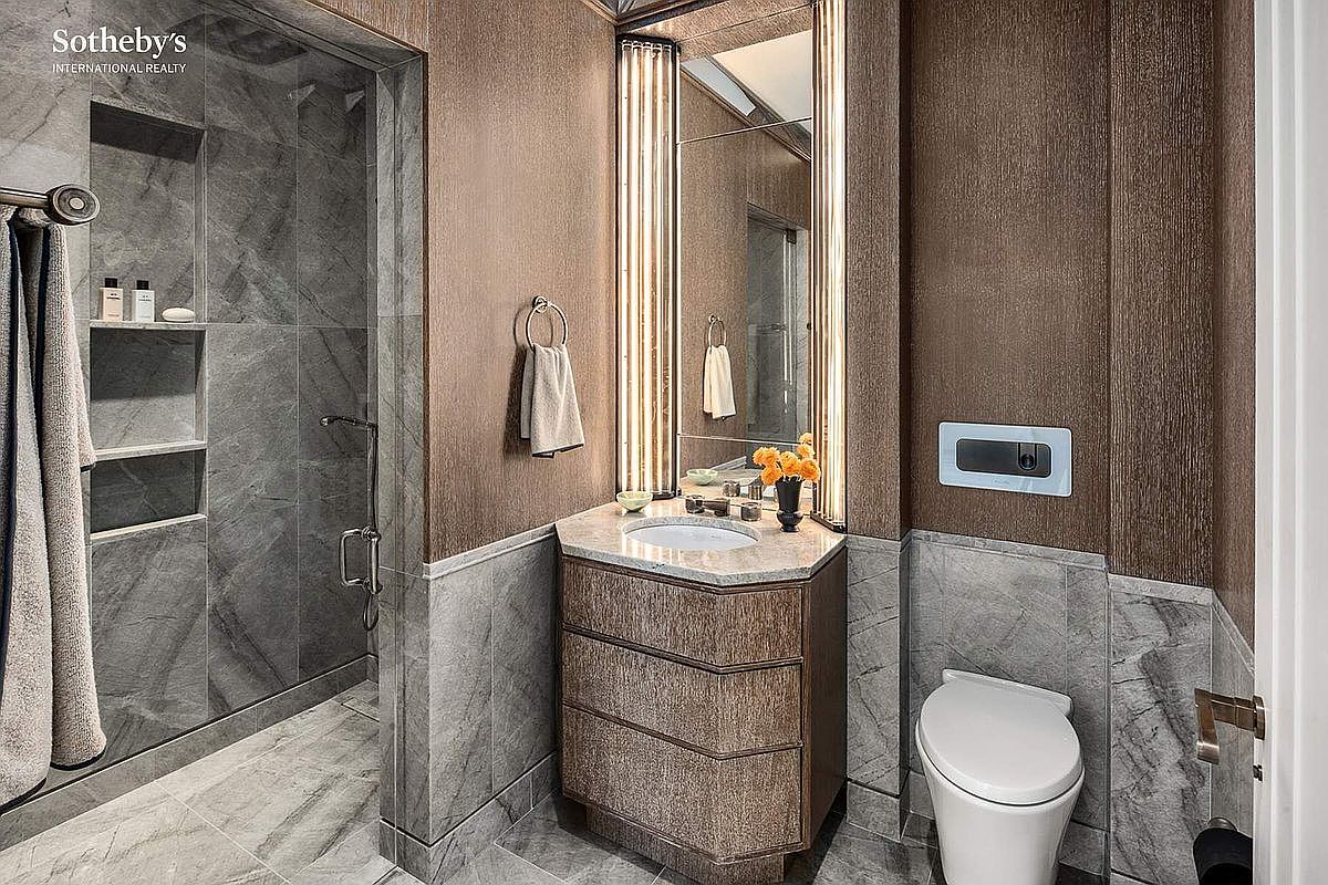 This sophisticated powder room features a striking blend of textured brown wallpaper and cool-toned grey marble tiling. The space is anchored by a custom vanity with a stone countertop and a sleek, wall-mounted toilet, complemented by a walk-in shower with integrated shelving. The vertical lighting flanking the mirror adds a touch of modern luxury, creating an elegant and polished atmosphere.
