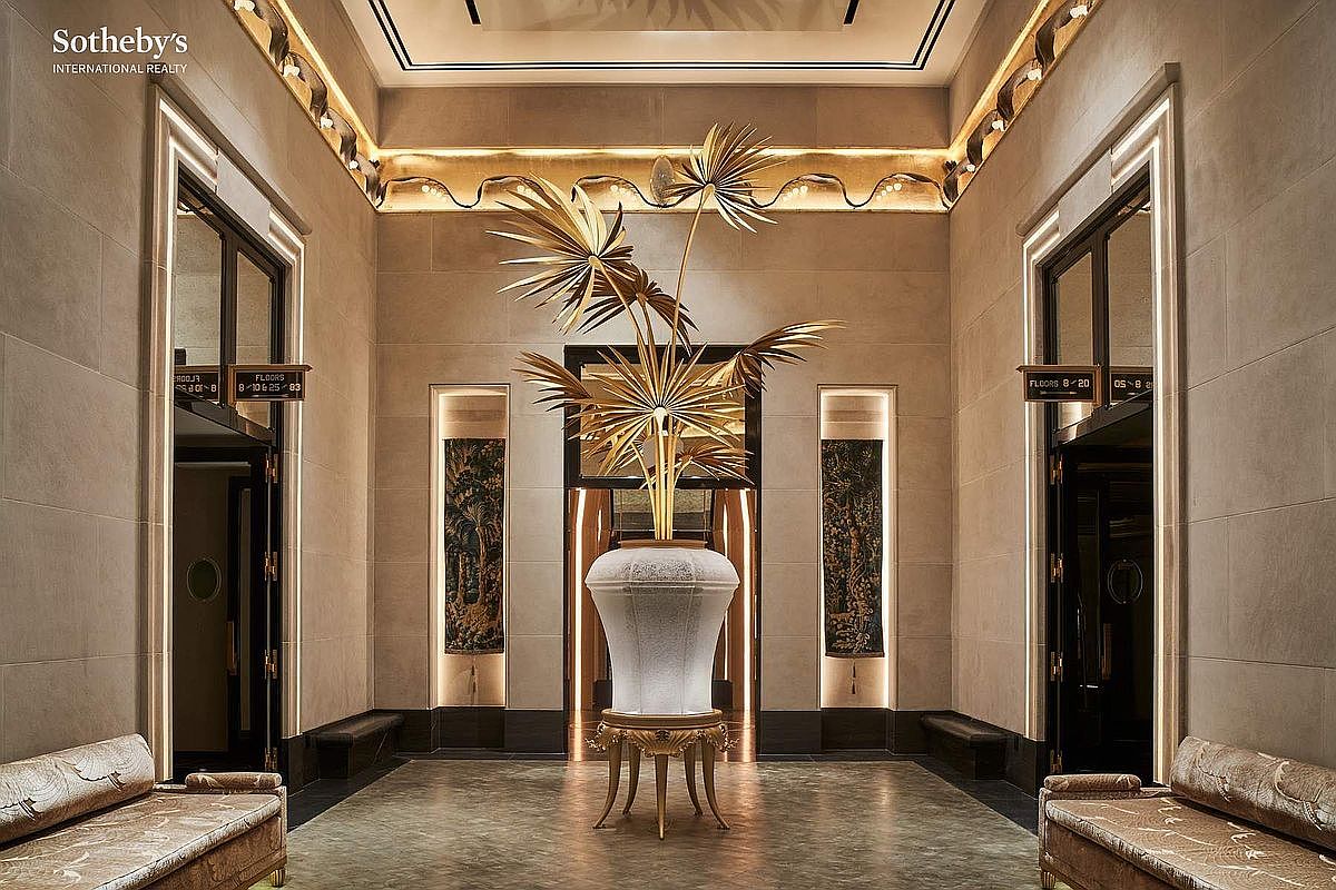 This grand, symmetrical lobby or elevator foyer exudes opulence with its high ceilings, warm ambient cove lighting, and sophisticated stone-clad walls. A striking, large-scale golden palm sculpture sits atop an ornate pedestal in the center, flanked by two elegant benches and framed by dark, metallic elevator doors. The perspective is perfectly centered and cinematic, emphasizing the luxurious, high-end architectural design and refined aesthetic of the building's common area.