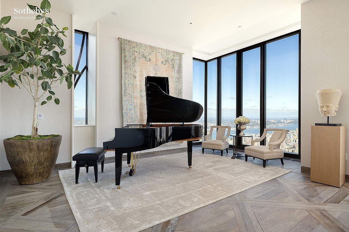 This sophisticated living space features a grand piano as its centerpiece, set against a backdrop of floor-to-ceiling windows that offer expansive city views. The room is elegantly appointed with a neutral-toned area rug, two plush armchairs, and a decorative bust on a pedestal, all set upon rich, herringbone-patterned hardwood floors. The overall aesthetic is one of refined luxury and modern elegance, perfectly suited for high-end urban living.