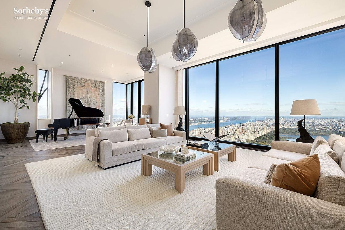 This expansive, high-end living room features floor-to-ceiling windows that offer a breathtaking, panoramic view of the city skyline and Central Park. The space is elegantly furnished with two neutral-toned sofas, a light wood coffee table, and a grand piano, all set upon a large, textured area rug. The modern aesthetic is complemented by unique, sculptural pendant lighting and a sophisticated, airy atmosphere that emphasizes luxury and scale.