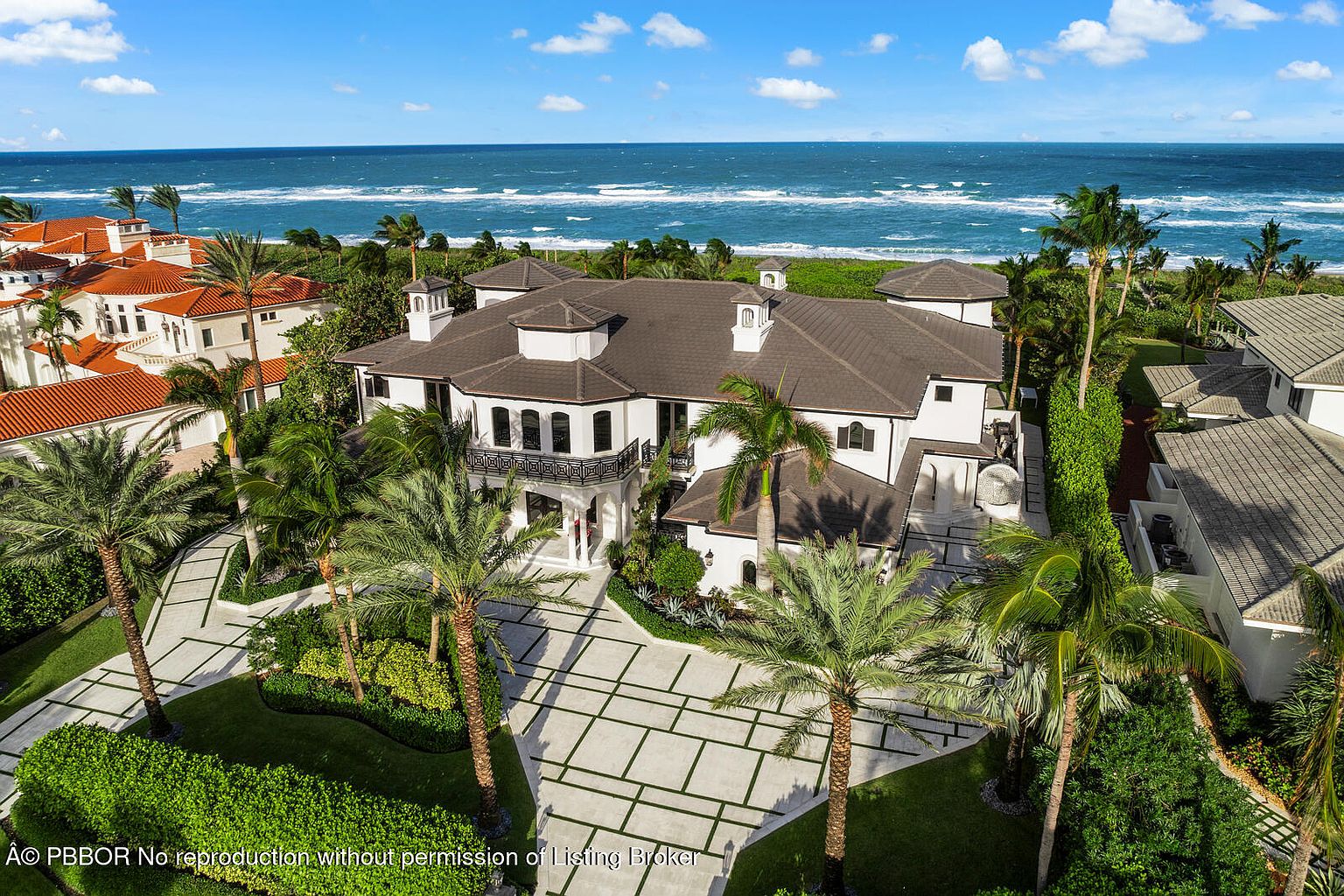 This aerial view showcases a luxurious beachfront estate with a grand driveway, meticulously landscaped grounds featuring palm trees and manicured lawns, and a stunning ocean backdrop. The architecture is elegant, with a dark roof contrasting against the white exterior, complemented by multiple balconies and arched windows, creating an impression of opulence and coastal living.