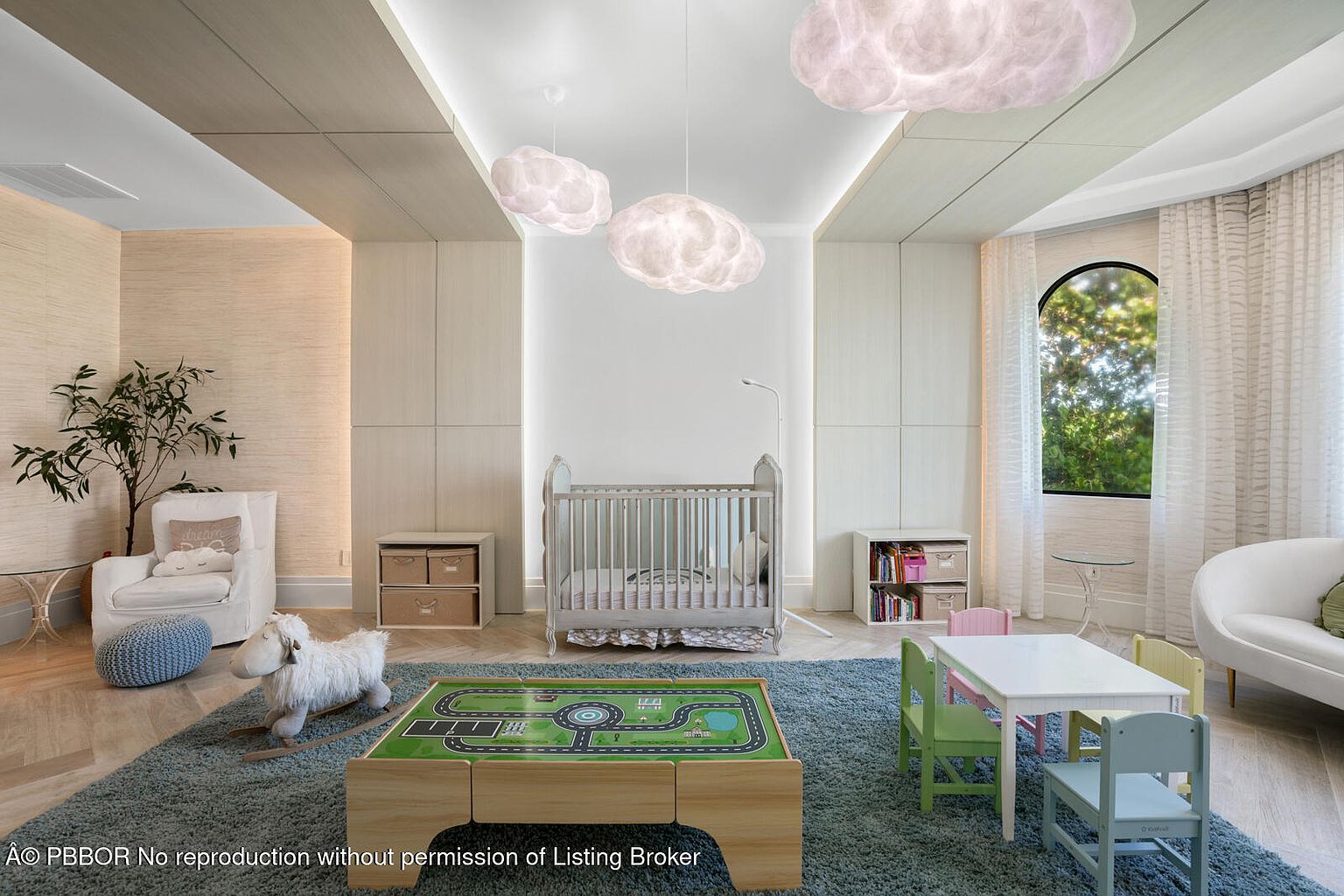 This is a charming children's bedroom featuring a crib, a play table with a race track design, and a small table with chairs. The room is decorated in soft, neutral tones with cloud-shaped light fixtures, creating a whimsical and calming atmosphere. A rocking sheep and a comfortable armchair add to the room's cozy and inviting feel.