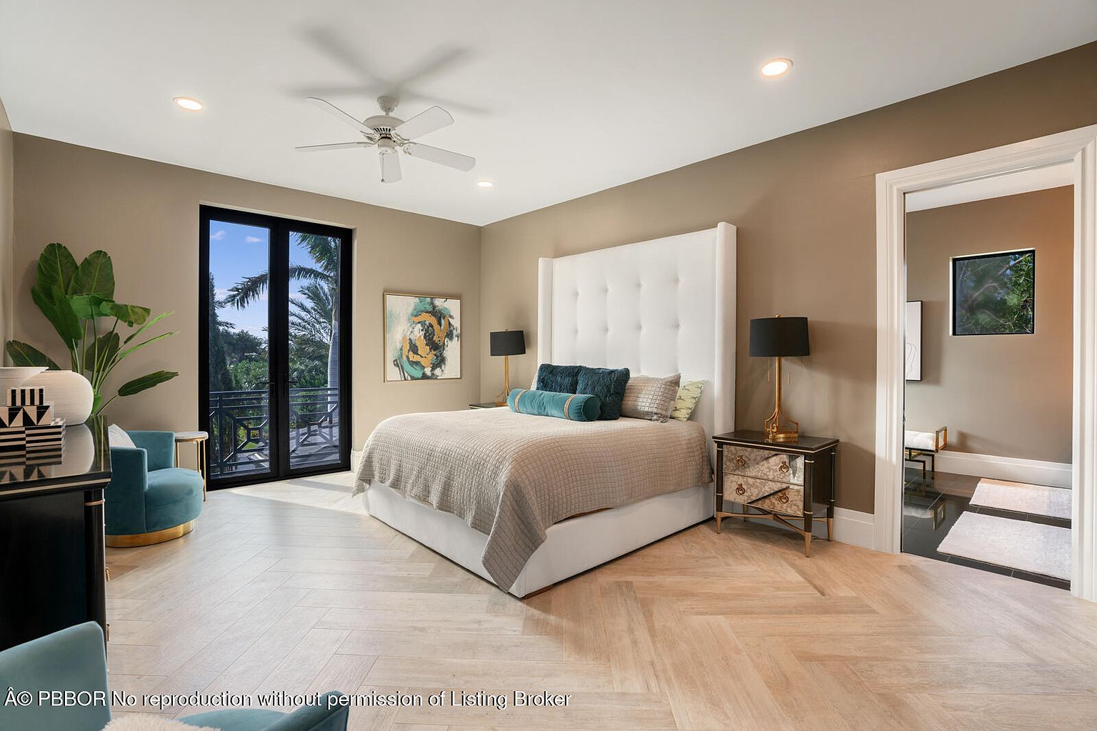 This is a primary bedroom featuring a large bed with a white tufted headboard, flanked by mirrored nightstands and lamps. The room has a neutral color palette with a herringbone wood floor and a door leading to an en-suite bathroom. A balcony door provides natural light and a view of the outdoors.
