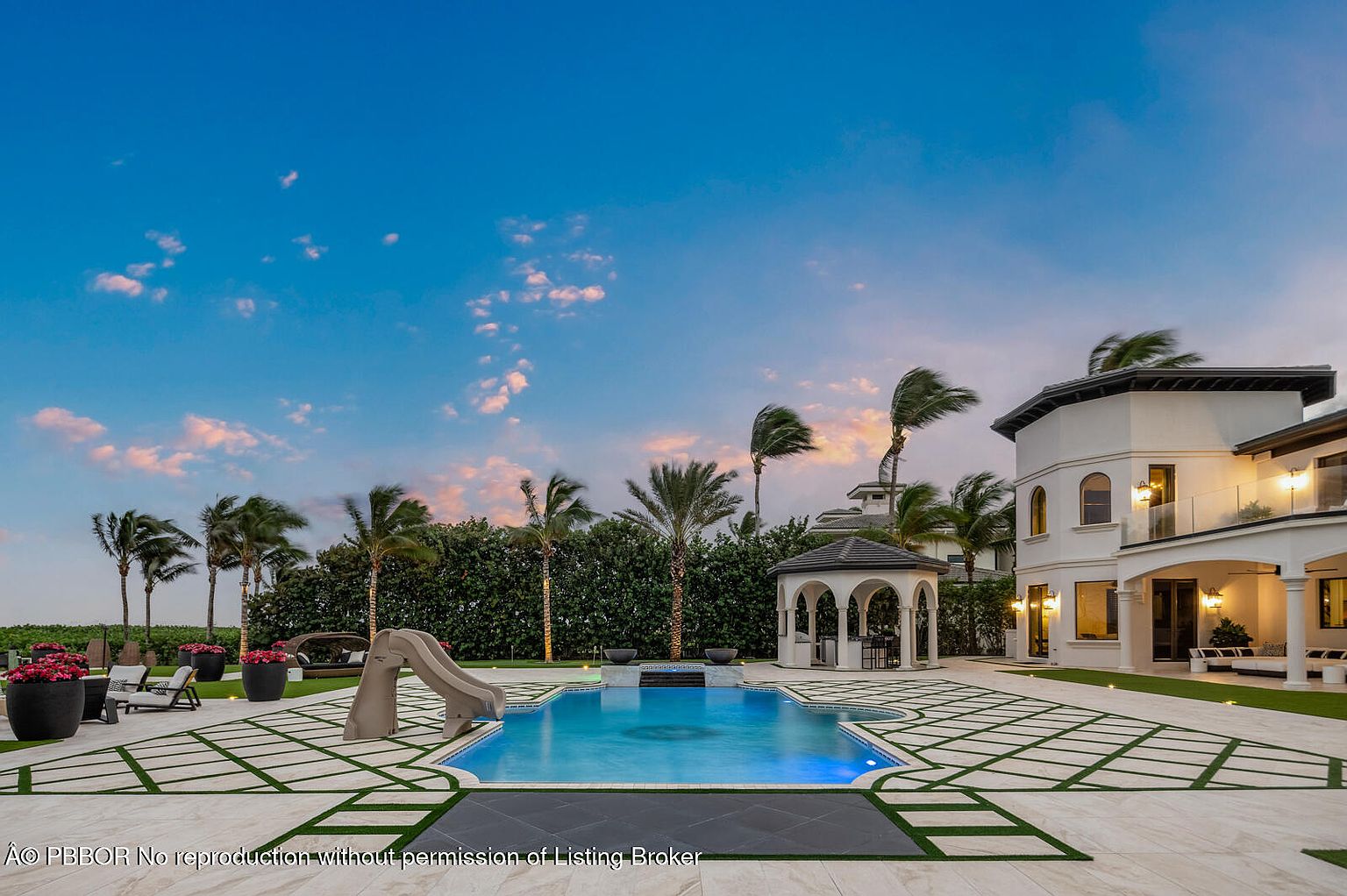 This image showcases a luxurious backyard featuring a stunning pool and spa area. The pool is surrounded by elegant stone tiling with grass accents, complemented by a slide and a charming gazebo. Lush landscaping and palm trees enhance the tropical ambiance, while the grand architecture of the house adds a touch of sophistication.