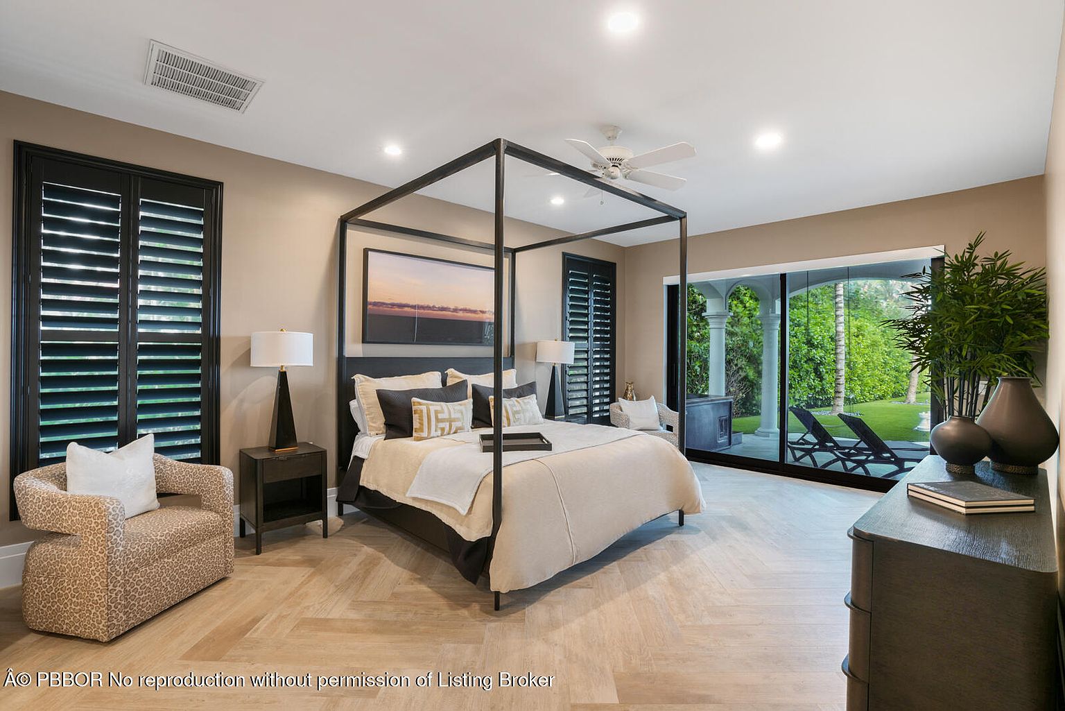 This is a luxurious primary bedroom featuring a four-poster bed with neutral bedding and decorative pillows. The room has a serene and sophisticated ambiance, enhanced by the natural light streaming through the large sliding glass doors that lead to an outdoor area. The herringbone wood flooring and elegant furnishings contribute to the room's upscale appeal.