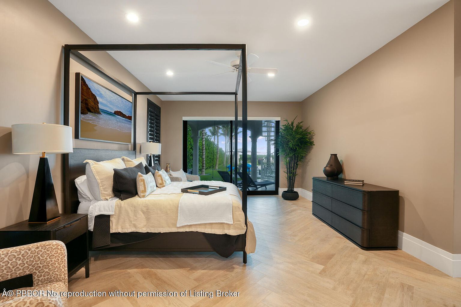 This is a primary bedroom featuring a four-poster bed with neutral-toned bedding and decorative pillows. A dark wood dresser sits against the wall, complemented by a potted plant. The room has a serene and luxurious feel, enhanced by the natural light filtering through the sliding glass doors leading to an outdoor area.