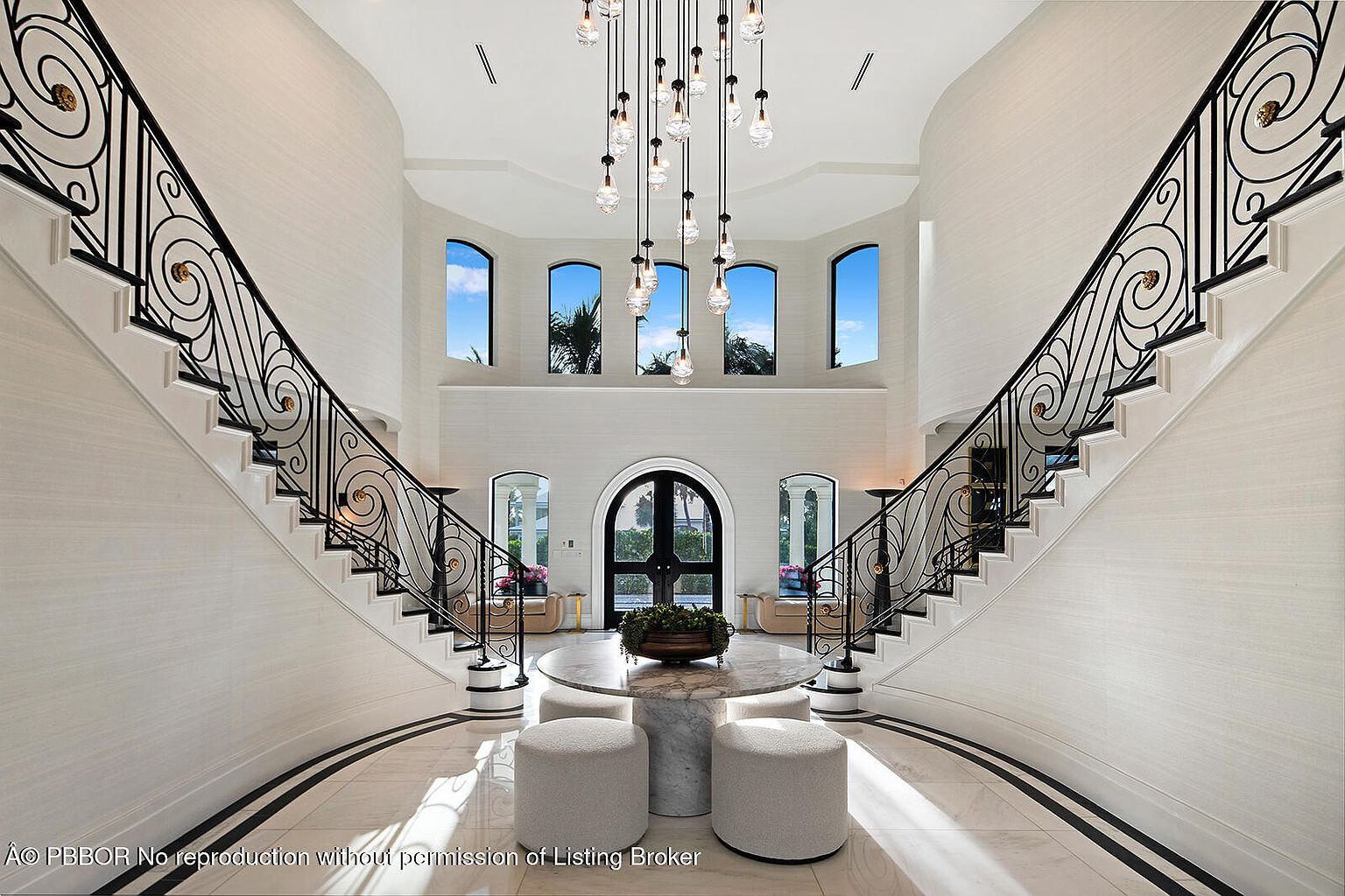 This grand foyer features dual staircases with ornate black iron railings and white risers, flanking a central seating area with a round marble table and upholstered ottomans. A striking chandelier hangs from the high ceiling, and arched windows provide natural light. The space is elegant and luxurious, creating a memorable first impression.