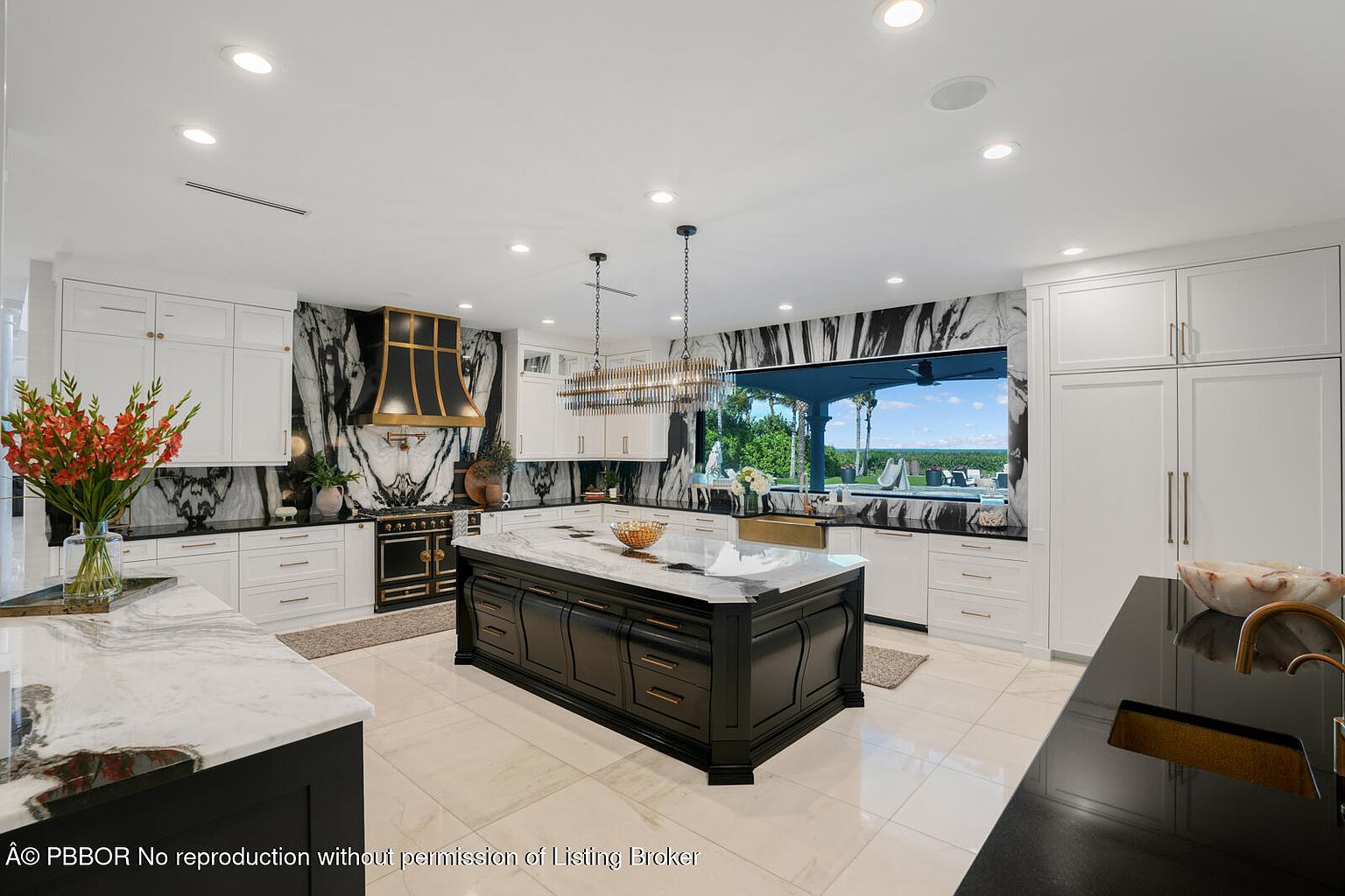 This is a spacious and luxurious kitchen featuring a large central island with a marble countertop and dark wood cabinetry. The kitchen is equipped with high-end appliances, including a black and gold range, and features white cabinetry along the walls. A large window provides a view of the outdoor pool area, and the overall design incorporates a modern aesthetic with contrasting dark and light elements.
