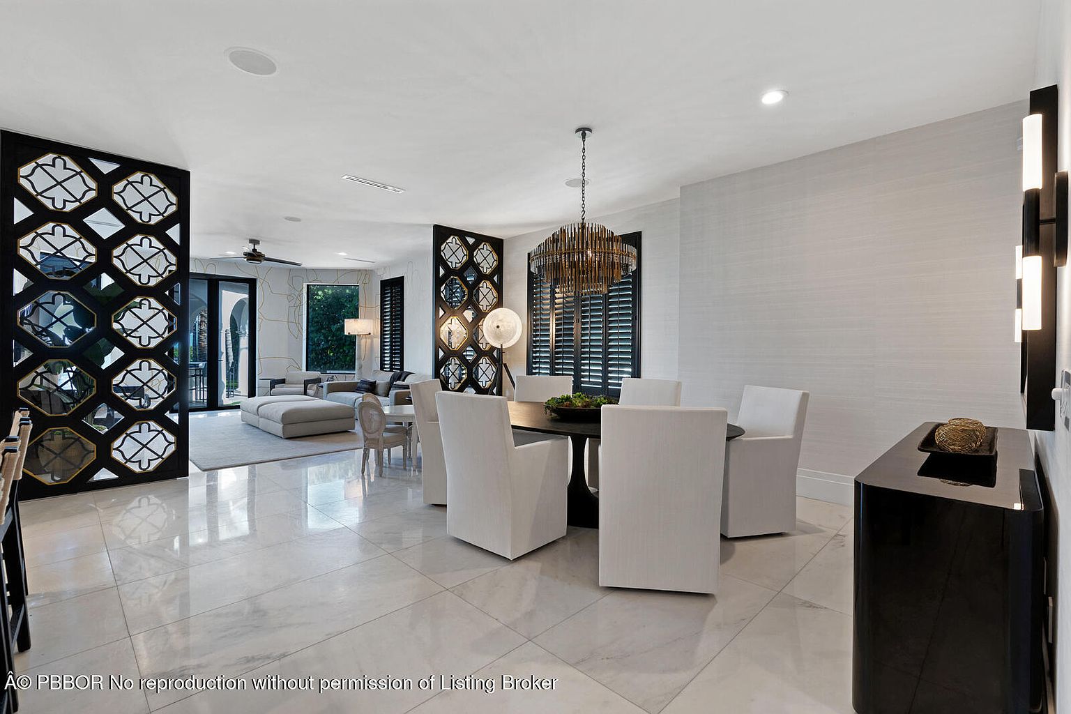 This interior shot showcases a modern dining room with a black round table surrounded by six white upholstered chairs. A striking chandelier hangs above the table, and a black console table with decorative items sits against the wall. The room features marble flooring and decorative screens, creating an elegant and sophisticated atmosphere.