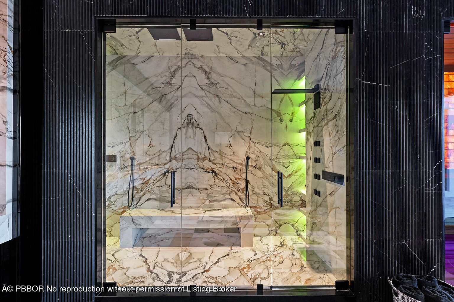 This image showcases a luxurious primary bathroom featuring a spacious walk-in shower with marble walls and a built-in bench. The shower is enclosed with glass and framed with black trim, and it includes multiple shower heads and body jets. Green ambient lighting adds a modern touch, creating a spa-like atmosphere.