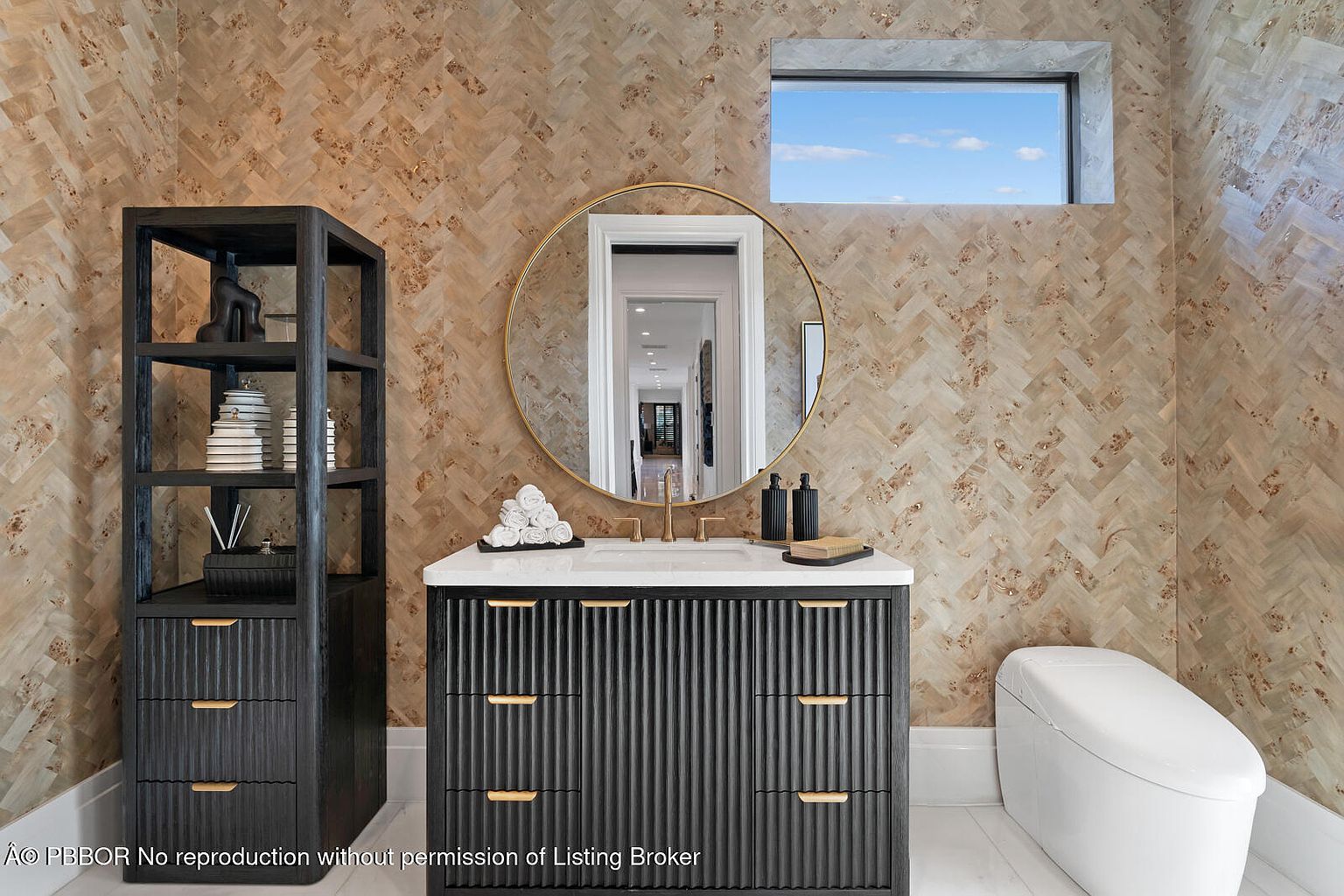 This is a stylish guest bathroom featuring a dark wood vanity with gold hardware and a white countertop. A round gold-framed mirror hangs above the sink, and a small window provides natural light. The walls are covered in a unique patterned wallpaper, and a matching dark wood shelving unit stands to the left of the vanity. A modern toilet is visible to the right. The perspective is a medium shot, showcasing the vanity and surrounding elements.