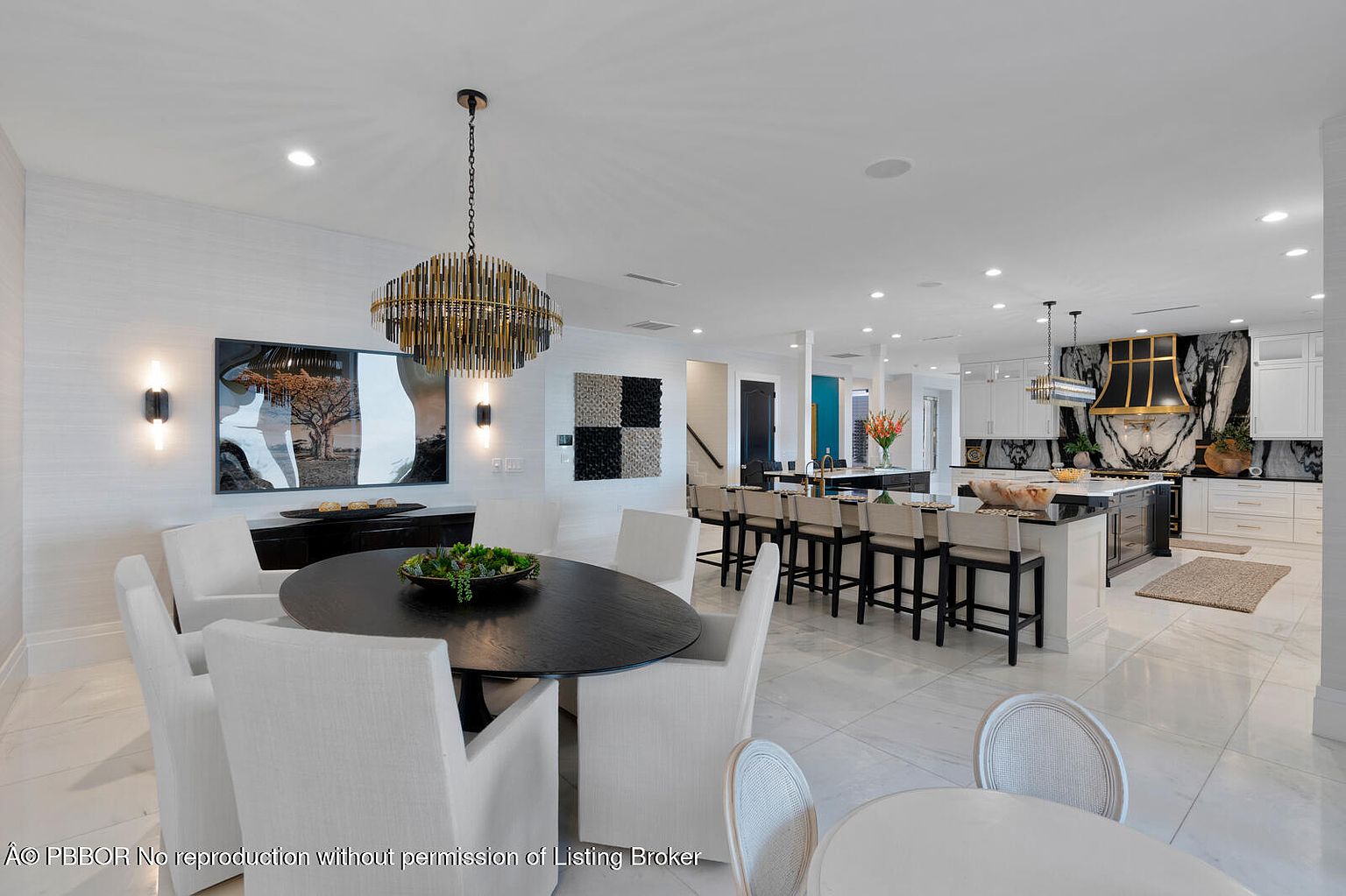 This interior shot showcases a modern dining area seamlessly integrated with a kitchen space. The dining area features a dark, round table surrounded by white upholstered chairs and an ornate chandelier. The kitchen boasts a large island with bar seating, black and white marble backsplash, and high-end appliances, creating a luxurious and open living space.