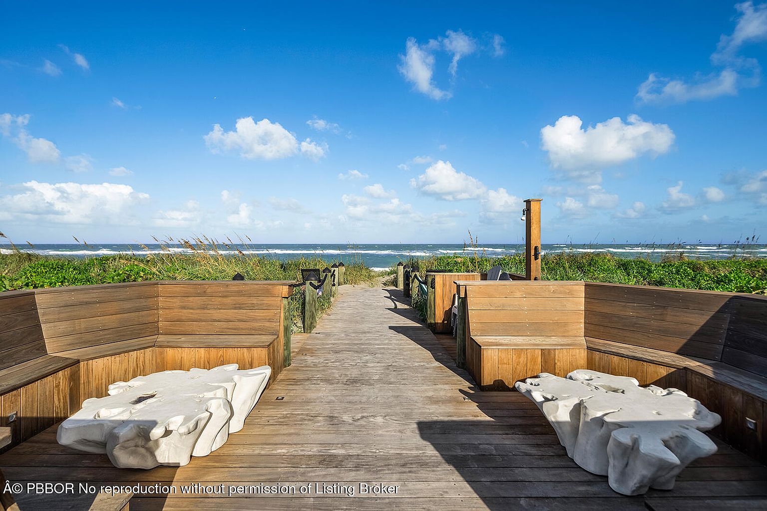 This image showcases a beautifully designed wooden deck or patio area leading towards the ocean. The deck features built-in wooden benches with unique, sculptural white tables, creating a relaxing and inviting outdoor space. The view extends to the ocean, framed by dune vegetation, offering a serene and private setting.