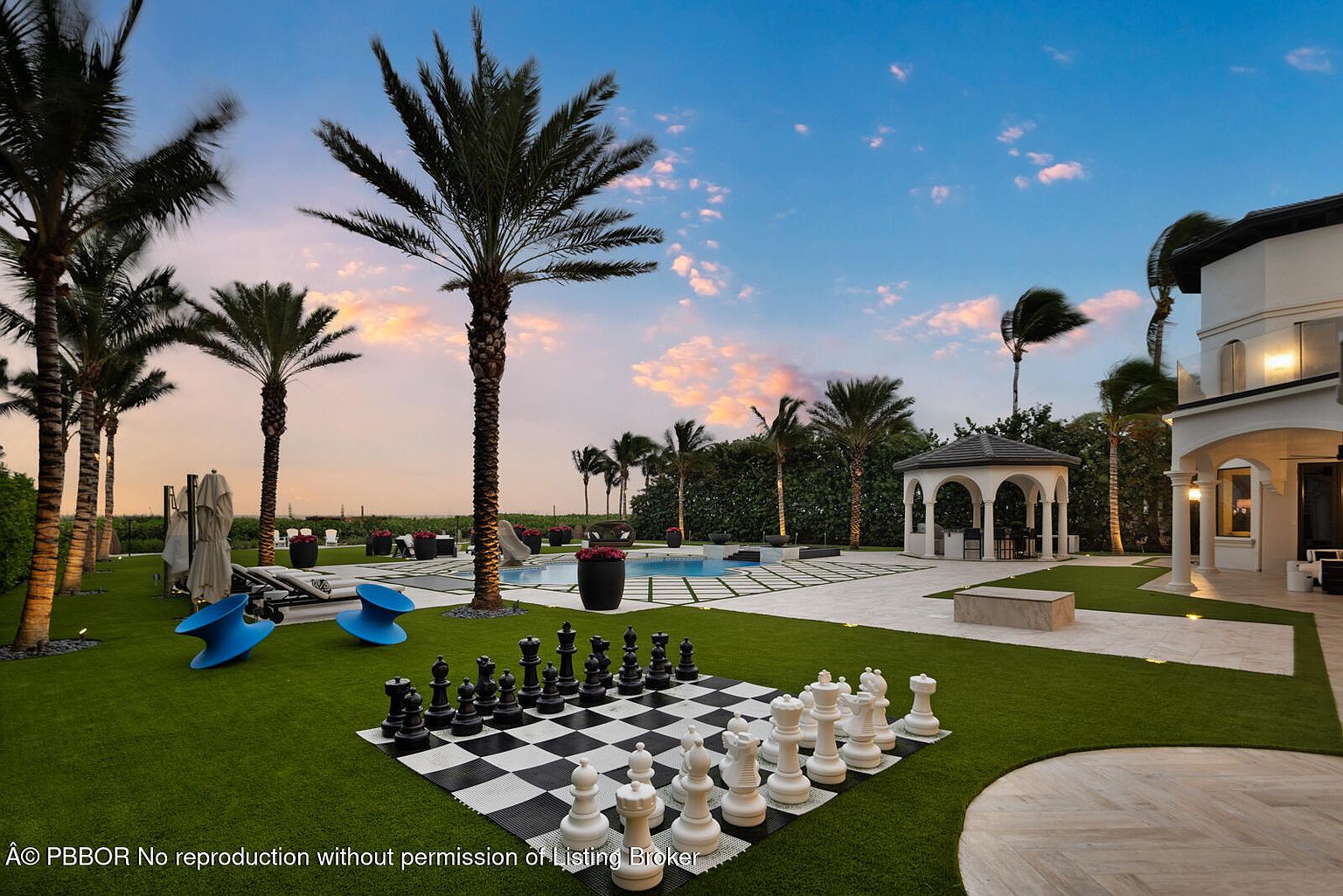 This image showcases a luxurious backyard featuring a large chessboard, a swimming pool, and a gazebo. Palm trees dot the landscape, adding a tropical feel, while the manicured lawn and elegant patio contribute to the property's high-end appeal. The scene is set against a beautiful sky, enhancing the overall impression of a serene and upscale outdoor living space.