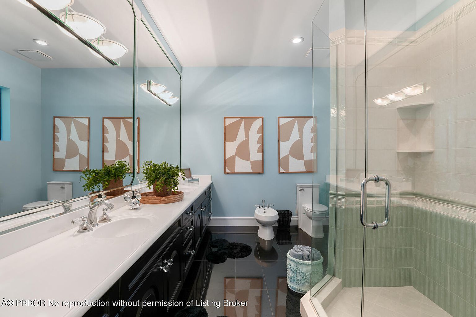 This is a well-lit bathroom featuring a double vanity with a white countertop and dark cabinetry. The walls are painted a light blue, complemented by framed abstract artwork. A glass-enclosed shower and a bidet are also visible, adding to the bathroom's functionality and style.