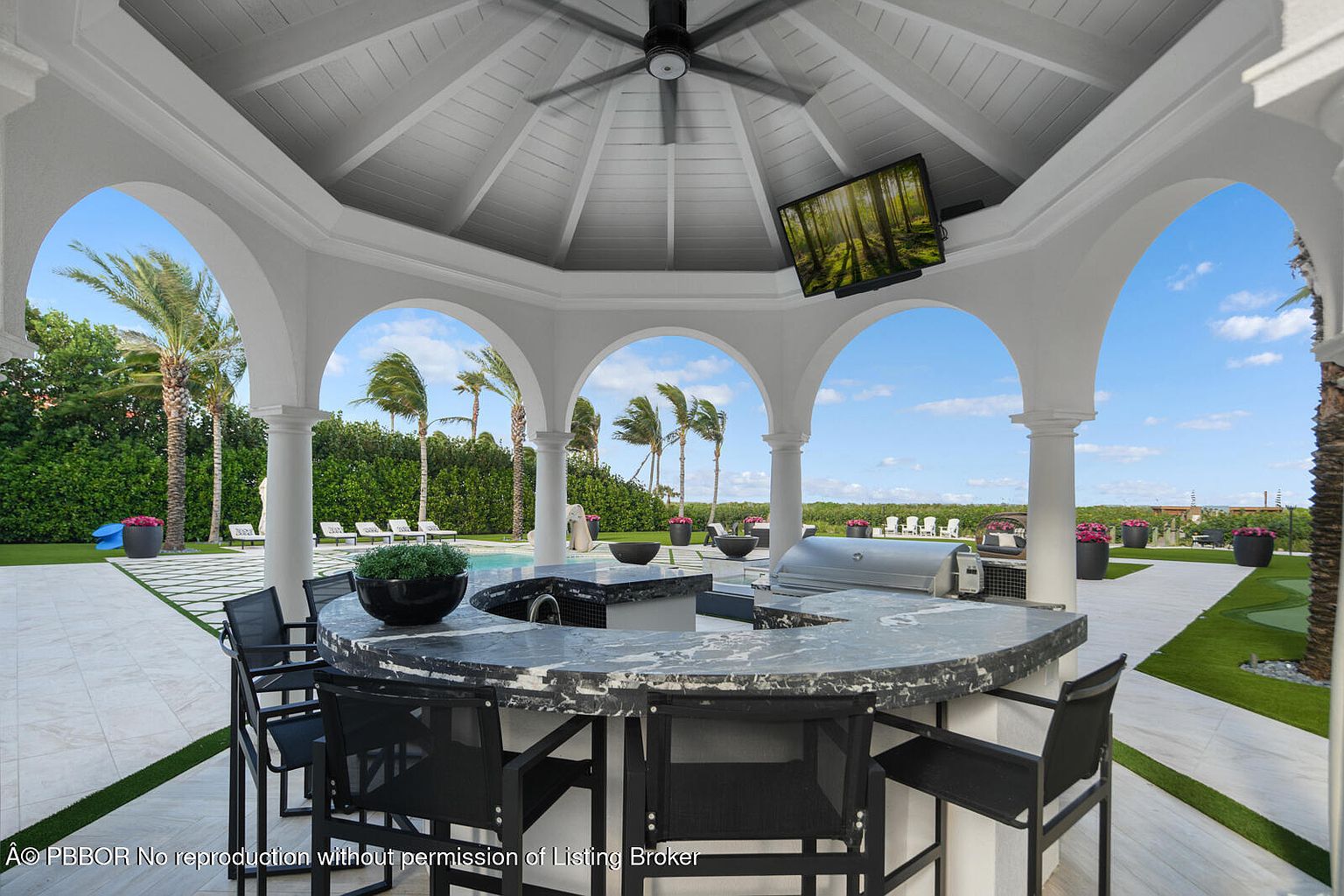 This image showcases an outdoor entertainment area featuring a covered patio with a built-in bar, complete with a marble countertop and bar seating. A large grill and outdoor television enhance the space, while the surrounding landscape includes palm trees, manicured lawns, and a glimpse of a pool area. The overall impression is one of luxury and outdoor living.