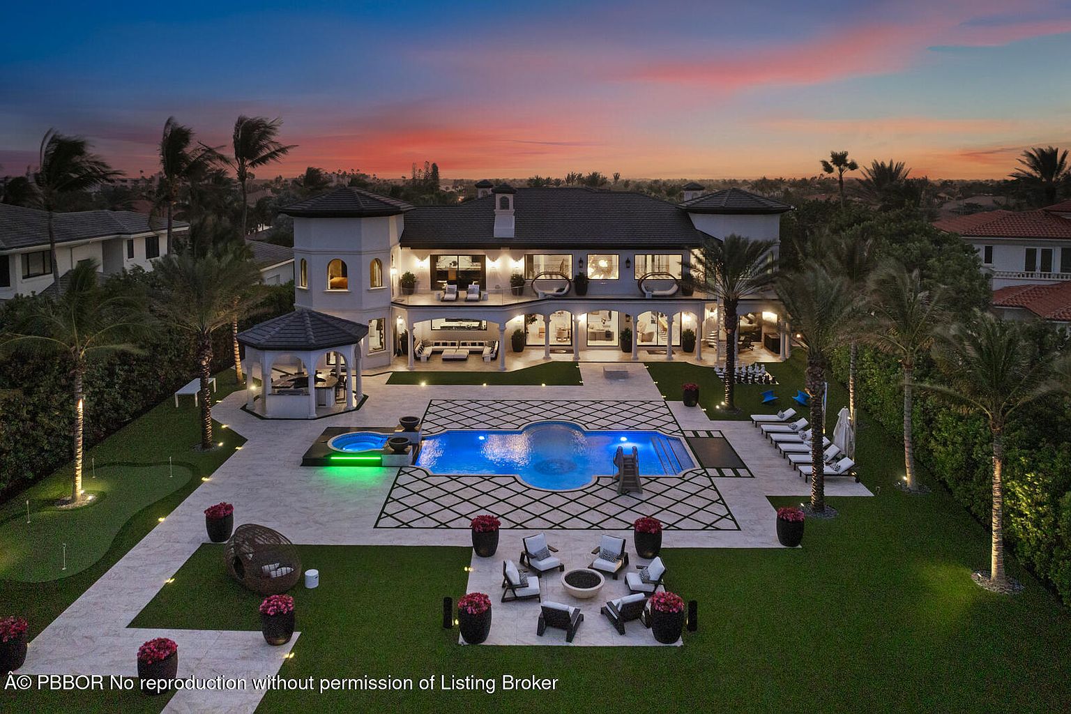 This aerial view showcases a luxurious estate at dusk, highlighting a large swimming pool with a spa, surrounded by a meticulously landscaped yard with palm trees and outdoor seating areas. The grand two-story residence features elegant architecture with multiple balconies and expansive windows, creating an inviting and upscale ambiance.