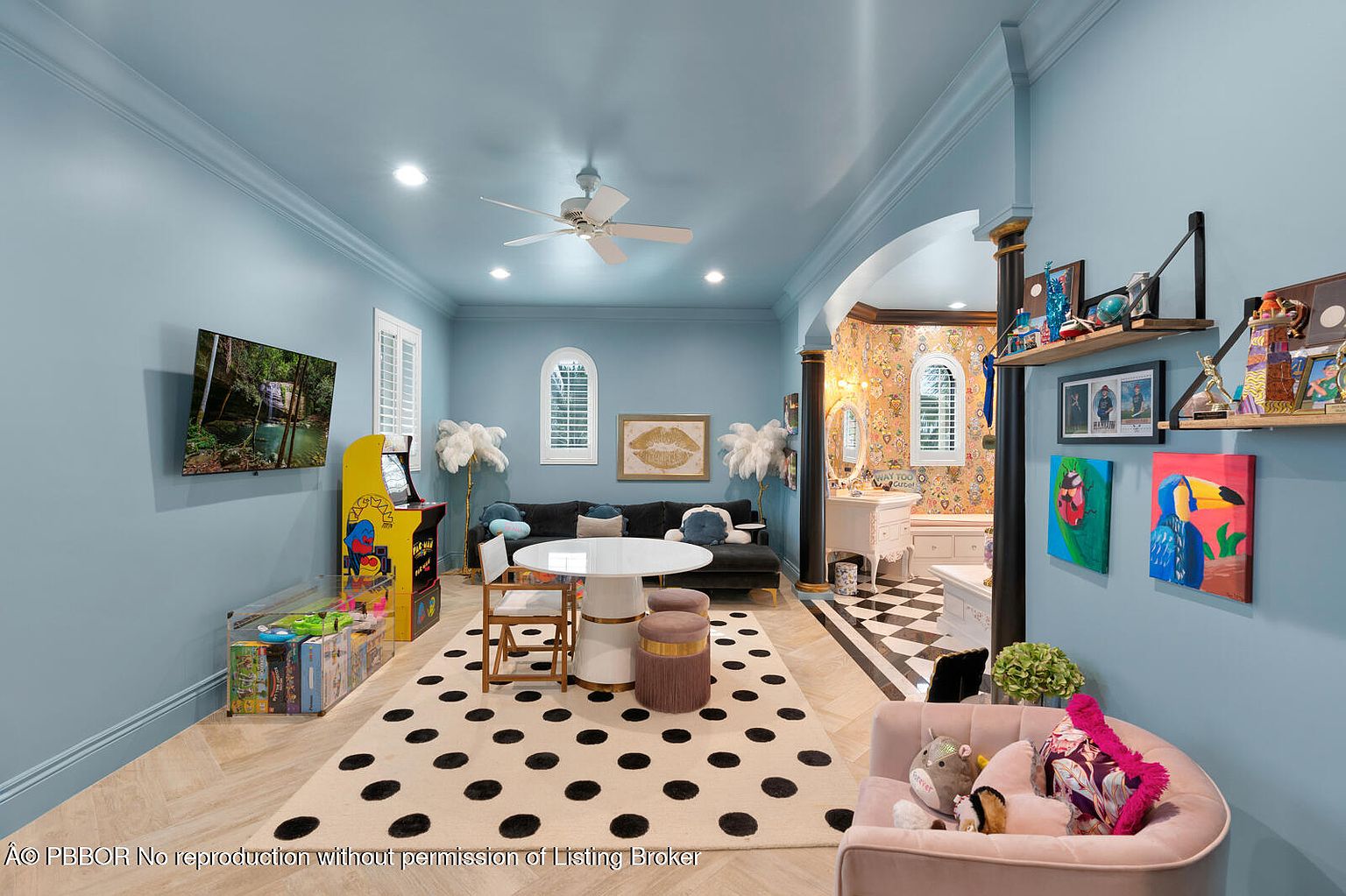 This vibrant living room features light blue walls, a black sofa with decorative pillows, and a round white table with chairs and stools. A polka dot rug adds a playful touch, while an arcade game and a TV provide entertainment. The room leads into a bathroom with patterned wallpaper and black and white checkered flooring, creating a unique and stylish space.